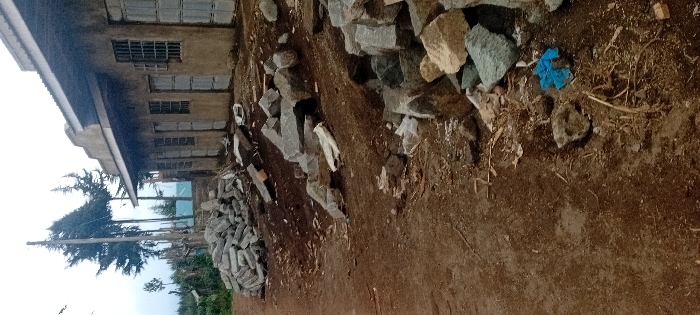 A building with barred windows sits beside piles of gray stones and construction debris. The ground is dirt, scattered with straw and a blue cloth. A stack of rectangular blocks rests at the edge of the frame, near a tree and some greenery. The photograph is taken at an angle, possibly with a wide-angle lens, resulting in some distortion.
