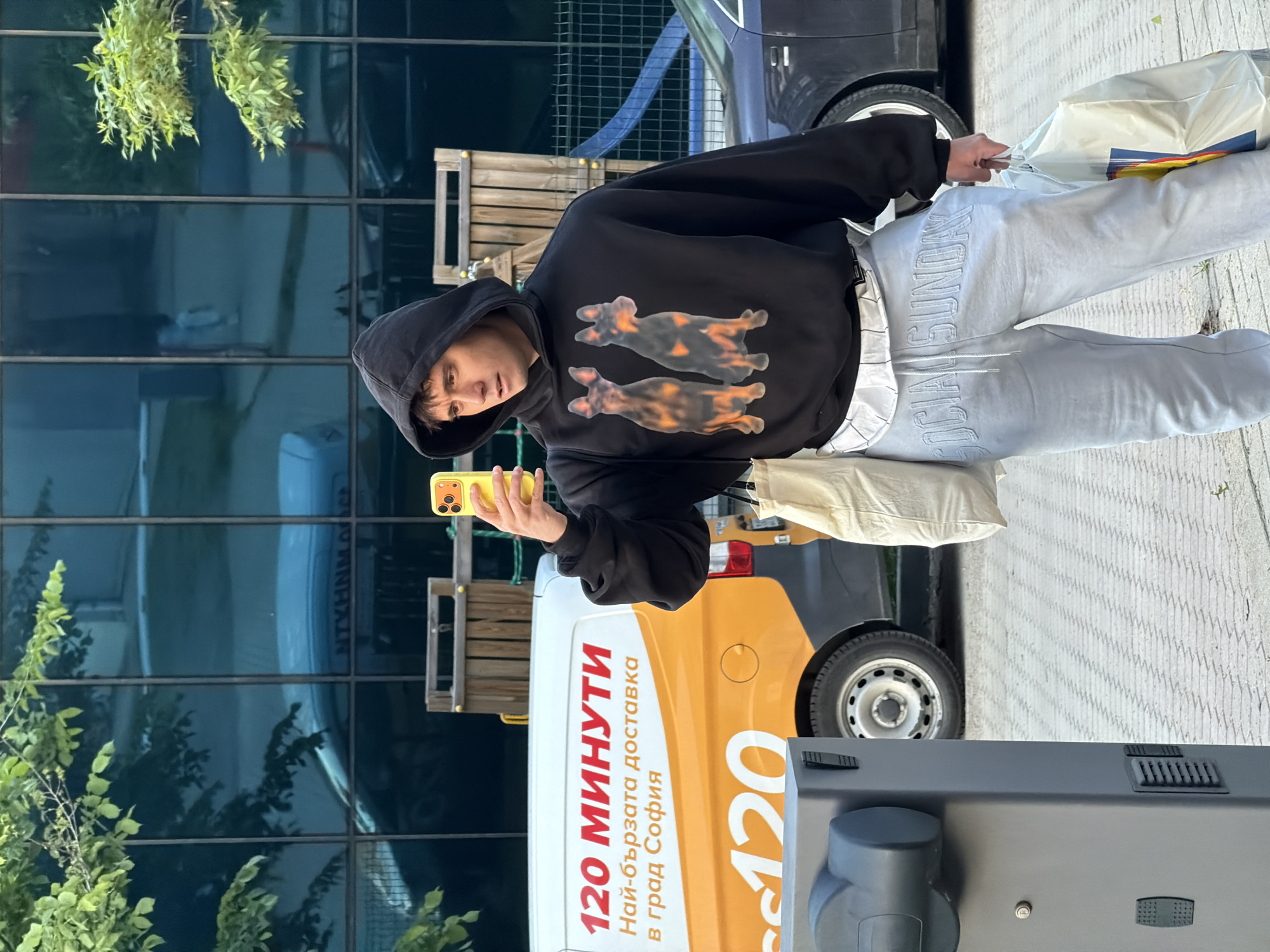 A young man in a black hooded sweatshirt with dog graphics and light grey sweatpants is holding a yellow smartphone while standing next to a white and orange delivery van. The van has "120 МИНУТИ Най-бързата доставка в град София" written on its side, indicating a delivery service within the city of Sofia, Bulgaria. The man is also carrying shopping bags. The setting appears to be an urban environment with a modern glass building visible in the background.