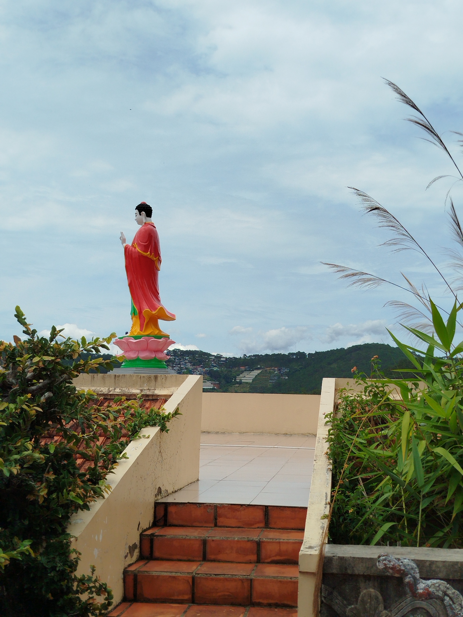 A statue of a figure, robed in red and standing on a pink lotus base, is on a roof. Steps lead up to the roof, which is tiled. Plants grow on either side of the steps. In the background is a landscape of hills and some buildings, under a cloudy sky. This location is likely in Da Lat, Vietnam.
