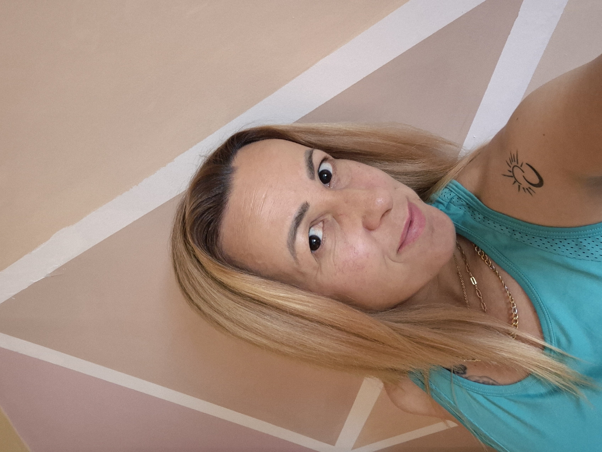 A woman with blonde hair and light skin is captured in a medium shot, looking towards the camera with a slight smile. She is wearing a turquoise tank top and two gold necklaces. On her left shoulder, she has a small black tattoo of a crescent moon with a sunburst. The background is a wall with a geometric pattern of diagonal lines in shades of beige, pink, and white. The lighting is even, suggesting an indoor setting.