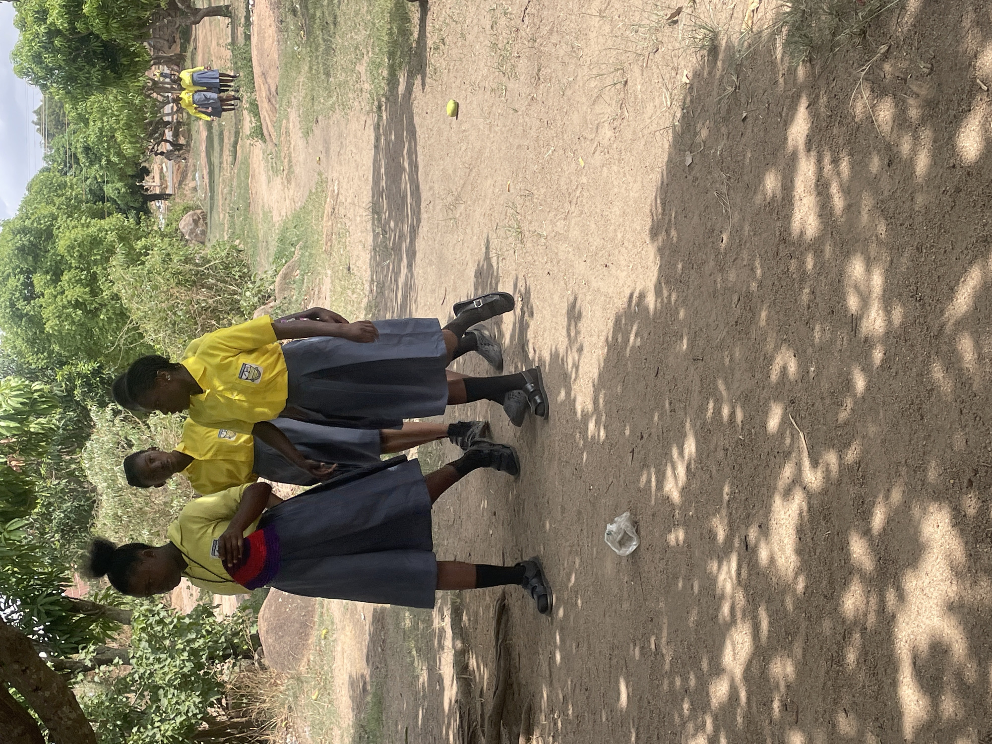The image depicts three young women in school uniforms standing on a dirt path. The uniforms consist of yellow shirts and grey skirts, paired with black socks and sandals. One of the women carries a colorful bag. The background shows a natural environment with trees and foliage. In the distance, there are additional figures, also possibly school children. The path is littered with a yellow fruit, a plastic bag, and shadows cast by the surrounding trees. The location is believed to be in Jos, Nigeria.
