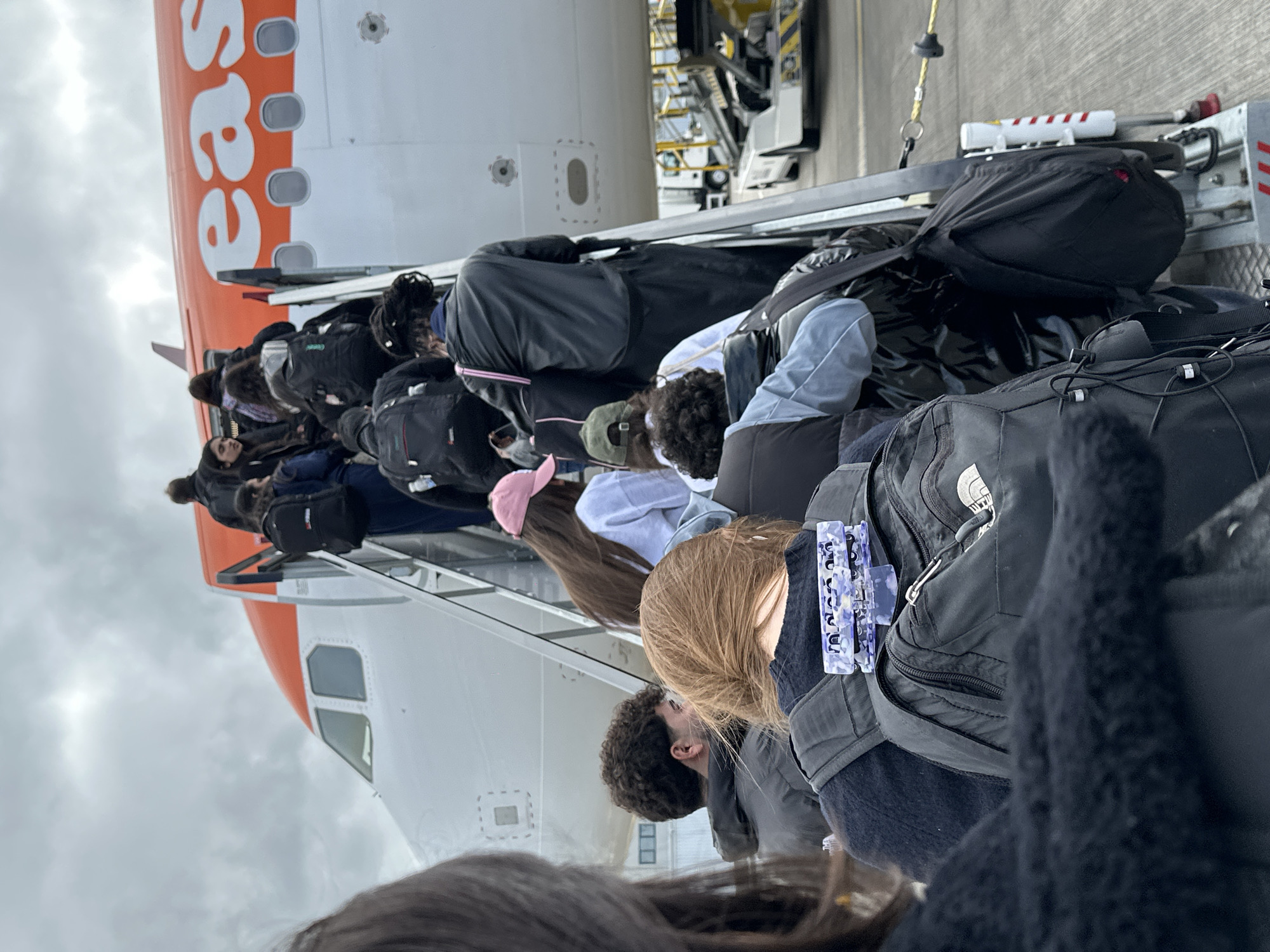 Passengers are boarding an EasyJet airplane. The airplane is white with an orange tail, emblazoned with the word "easyJet." People are walking up a mobile staircase directly into the plane, each carrying a backpack or bag. The scene suggests the exterior boarding procedure common at smaller airports. The location is presumably Southend Airport in Rochford, United Kingdom.