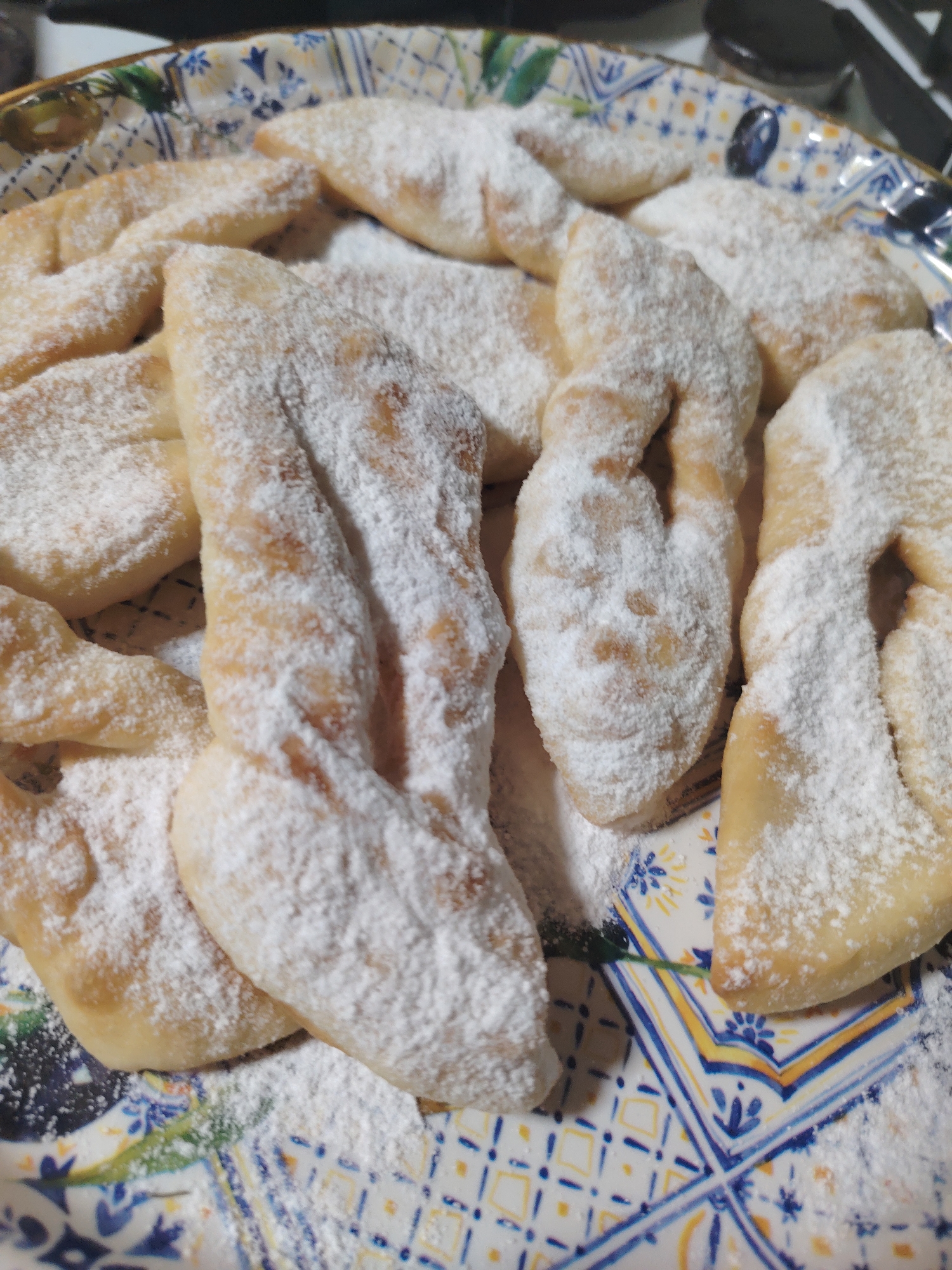A close-up shot features a pile of freshly made angel wings, a traditional Russian pastry, dusted generously with powdered sugar. The pastries are golden brown with a slightly puffed, irregular shape, showcasing the characteristic knot or twist in the center. They are arranged on a decorative platter with a blue, yellow, and white pattern, suggesting a home setting. The lighting is soft, indicating it might be daytime indoors. The overall impression is one of a sweet, homemade treat ready to be enjoyed. There is no visible text in the image.