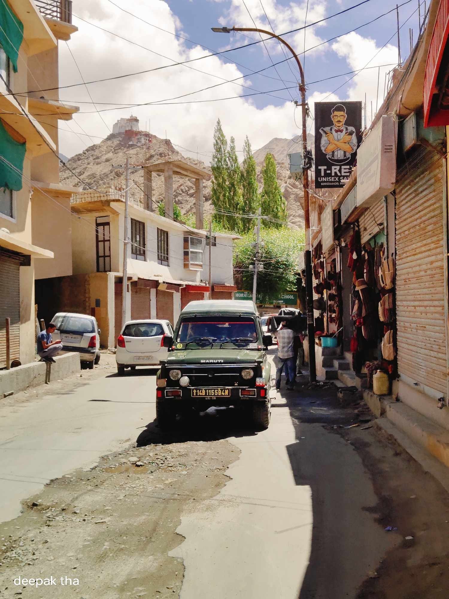 In the city of Leh, India, a street scene unfolds with a prominent green Maruti Gypsy vehicle in the foreground, its license plate reading "TJAB 115594N". To the right, a row of shops line the street, their metallic shutters closed. Bags and other items are displayed outside one shop. A man in a plaid shirt and jeans walks past, carrying a large object on his shoulder. Further down the street, a billboard advertises "T-Rex Unisex Salon". On the left side of the street, a man is seated on a low wall, looking at his phone. Behind him, a white car is parked. In the distance, a large monastery complex sits atop a rocky mountain under a cloudy sky. The road surface is uneven with patches of gravel and water.