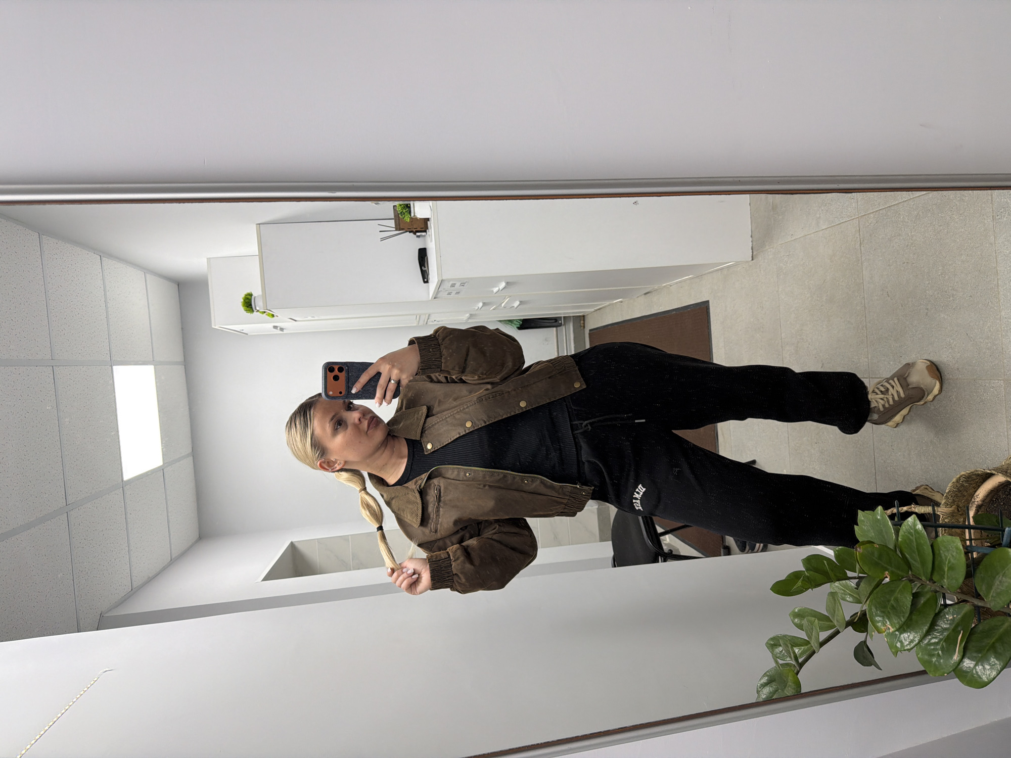 A young woman is seen in a mirror selfie, dressed in black sweatpants and a brown jacket over a black top. She has blonde hair styled in two pigtails and is holding one of them. She is holding a smartphone in her right hand to take the photo. The reflection shows a bathroom interior with light-colored tiles, a white cabinet, and a plant visible in the corner. The location is not specified in the image.