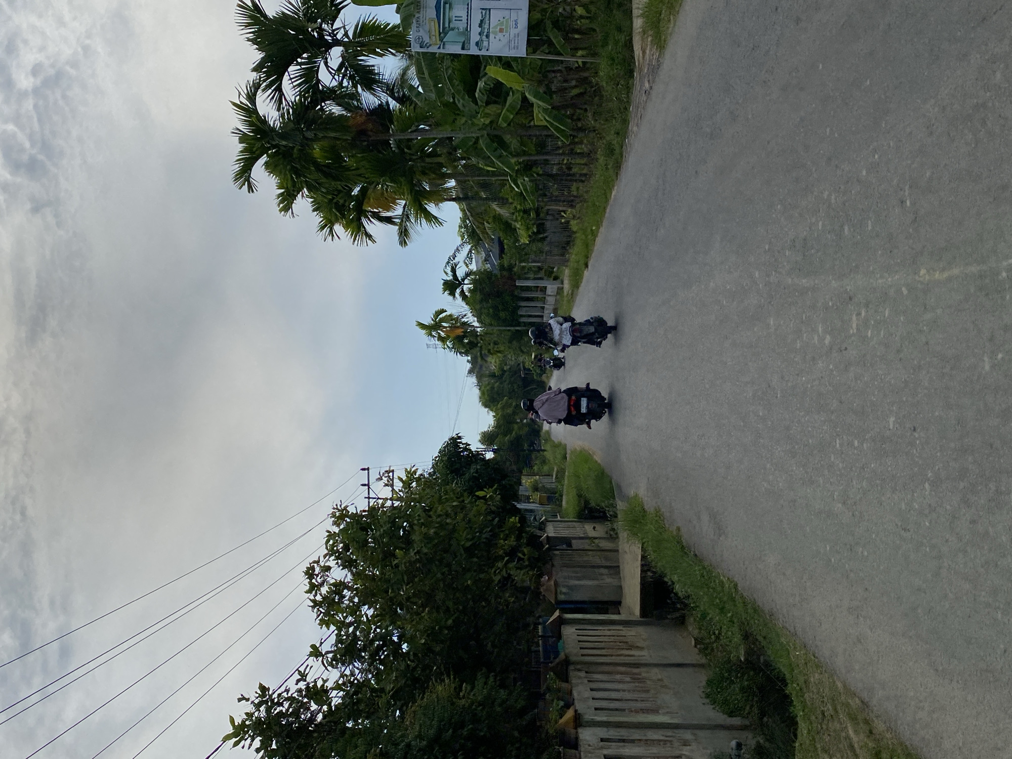The image captures a road scene in Lhokseumawe, Indonesia, likely on a cloudy day. Two individuals are riding motorcycles on a paved road that appears to be a rural or semi-urban area. The road is flanked by vegetation, including trees and palm trees. On the left side of the road, there is a low concrete wall with some plants growing alongside it, and further back, dense green foliage. On the right side, more palm trees and banana trees are visible, along with a billboard advertising a housing project. Power lines are strung across the sky, supported by utility poles. The sky is overcast with visible clouds. The perspective is from behind the motorcycles, suggesting the photo was taken while also moving on a motorcycle.