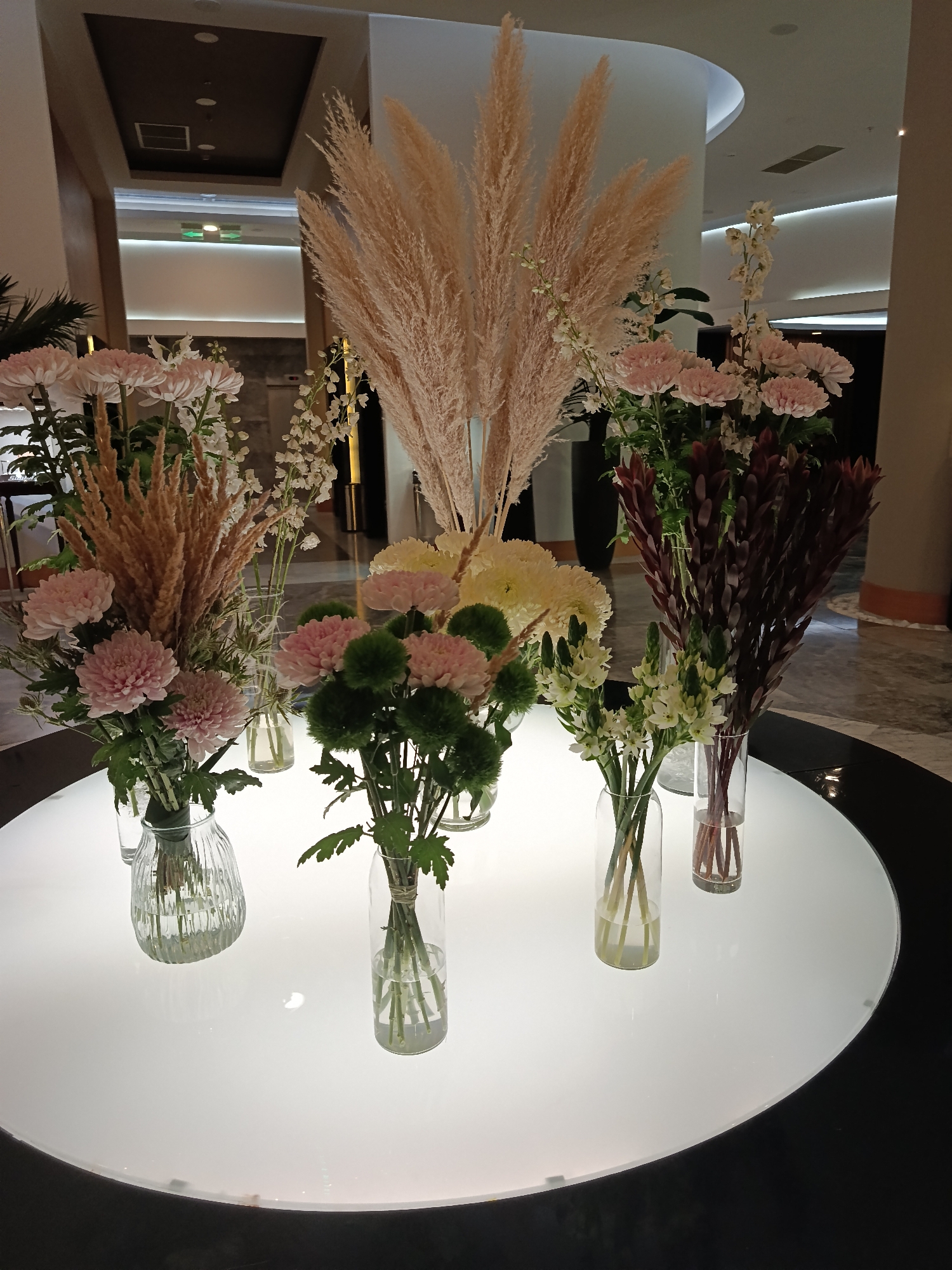 This image shows a floral arrangement on a round, illuminated table. The arrangement features several types of flowers, including pink chrysanthemums, white delicate flowers, and tall, feathery wheat-like stalks. The flowers are displayed in clear glass vases of various shapes and sizes. The table is set against a backdrop that appears to be an indoor space, possibly a hotel lobby or event venue, with modern architectural elements like recessed lighting and columns. There are no people or animals present in the image. The overall impression is one of sophisticated and elegant decoration. The location is identified as Sirius, Russia.