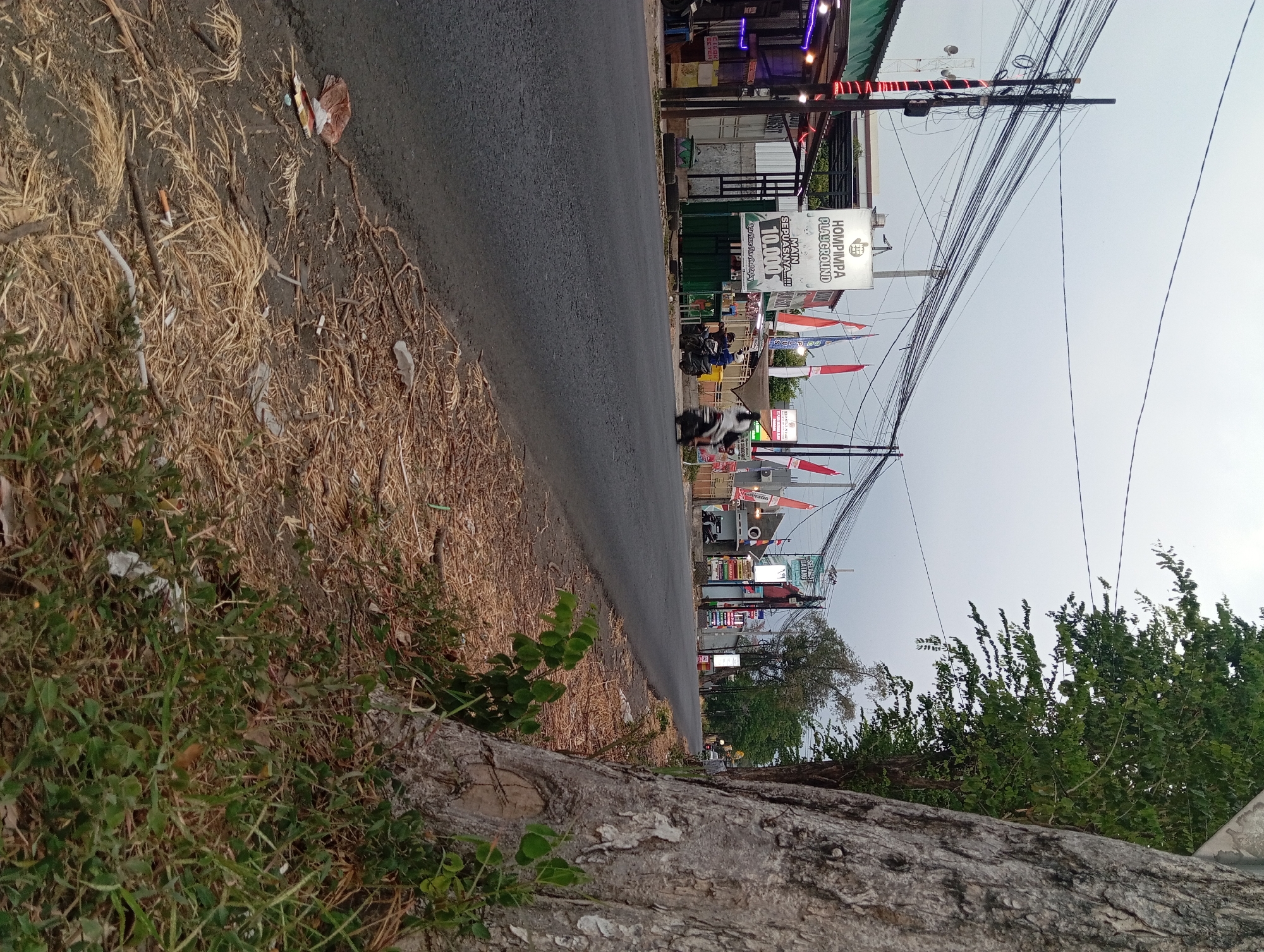 The image captures a street scene in Kebon Waris, Indonesia. The foreground features a large tree trunk on the right side, with dry grass and scattered debris along the roadside. A paved road occupies the middle ground, with several motorcycles visible, some in motion. To the left of the road, a row of buildings with awnings and signs line the street. One prominent sign displays "HOMPIMPA PLAYGROUND" with Indonesian text. Red and white flags are seen flying along the street. Power lines are strung overhead, supported by utility poles. The sky is overcast. The overall impression is that of a moderately busy street in a commercial area, with evidence of both activity and neglect due to the scattered litter.