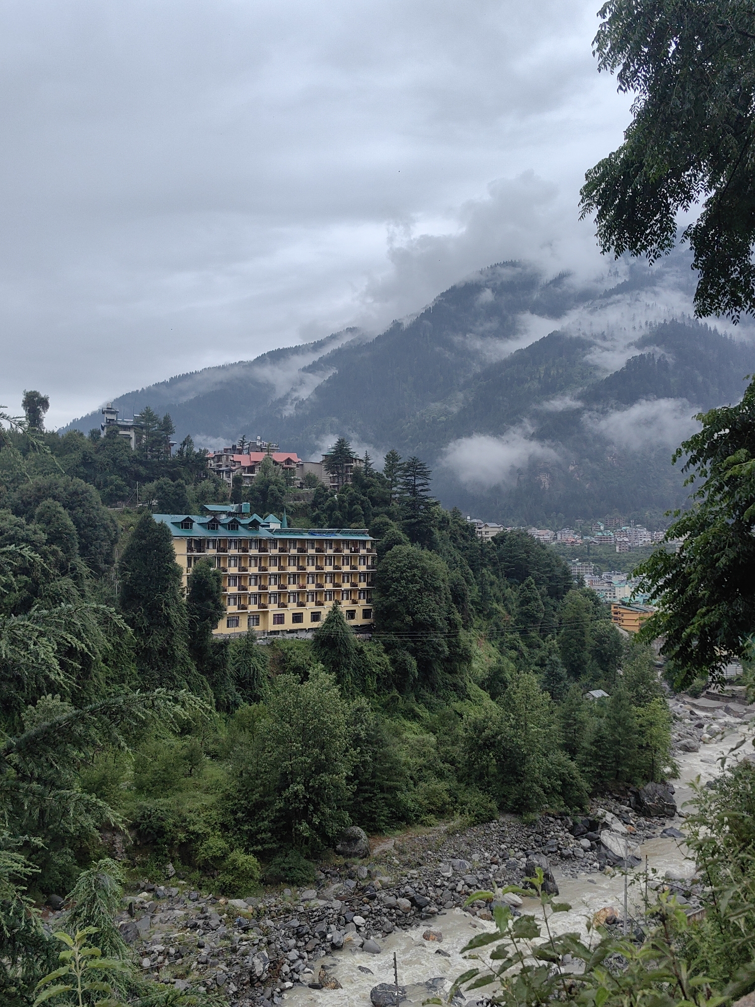 This image depicts a landscape in Aleo, India, characterized by a hotel situated on a verdant hillside overlooking a rocky river. The hotel, a prominent structure with a yellow facade and a green roof, features numerous balconies. It is nestled amongst dense evergreen trees. In the background, mist-shrouded mountains rise, their slopes covered in trees. The river, appearing somewhat turbulent and murky, flows over a bed of stones and rocks. The sky is overcast, contributing to a subdued atmosphere. Several other buildings are visible on the slopes and in the distance, suggesting a populated area. There are no identifiable people or specific actions occurring within the frame.