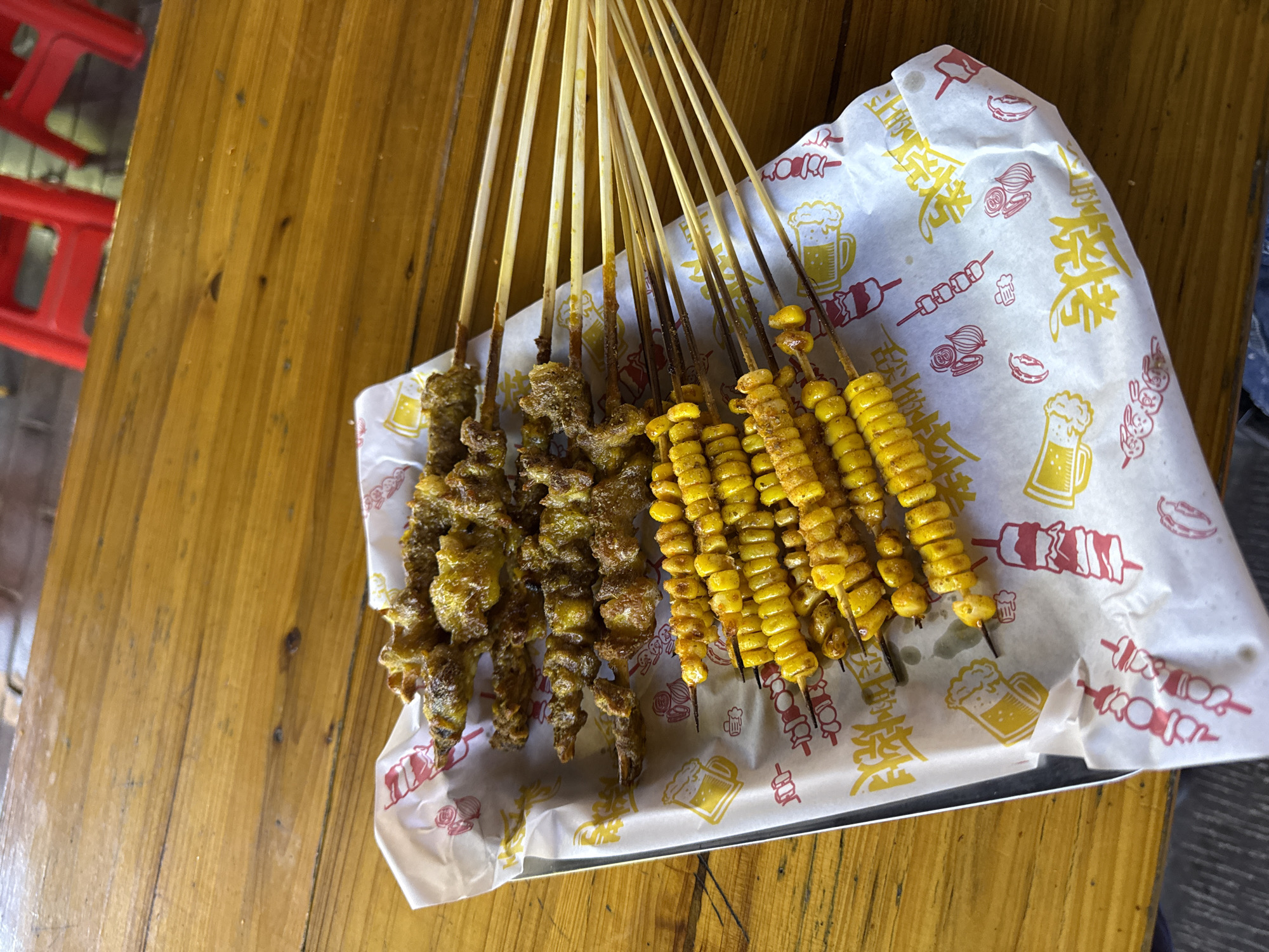 A top-down shot shows two groups of skewers, one of meat and one of corn, arranged on a white and yellow patterned paper on a metal tray, set on a wooden table in Wulingyuan, China. The meat skewers are on the left and the corn skewers on the right. The paper has illustrations of beer mugs, meat skewers, onions, and stylized Chinese characters. The skewers themselves are held upright with the wooden sticks extending upwards. A red plastic stool is visible in the background, indicating an outdoor or casual dining environment.