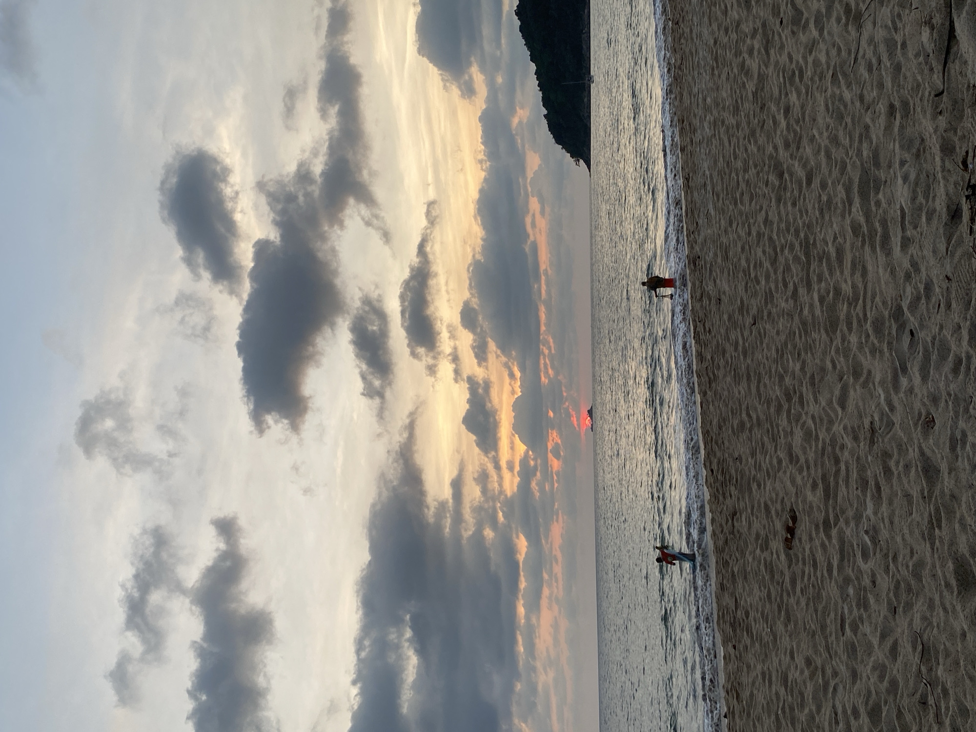 The image captures a picturesque beach scene at either sunset or sunrise in Choeng Thale, Thailand. The sky is a dominant feature, filled with a dramatic interplay of dark, voluminous grey clouds and bright, illuminated clouds tinged with golden, orange, and light blue hues. The sun, low on the horizon, casts a warm glow, with a distinct reddish-orange spot visible directly on the tranquil water surface.
The calm sea gently laps against the shore, reflecting the sky's light. A wide expanse of sandy beach with intricate ripple patterns stretches across the foreground. To the right, a dark, verdant mountain or hill rises, partially framing the celestial display.
Two individuals are visible in the shallow waters along the shoreline, silhouetted against the bright horizon. One person, wearing a red top, stands closer to the mountain. Further to the left, another person in a red top and blue shorts stands in the water, both seemingly observing the horizon. The scene conveys a sense of peaceful beauty typical of a tropical coastline at twilight. No text is visible.