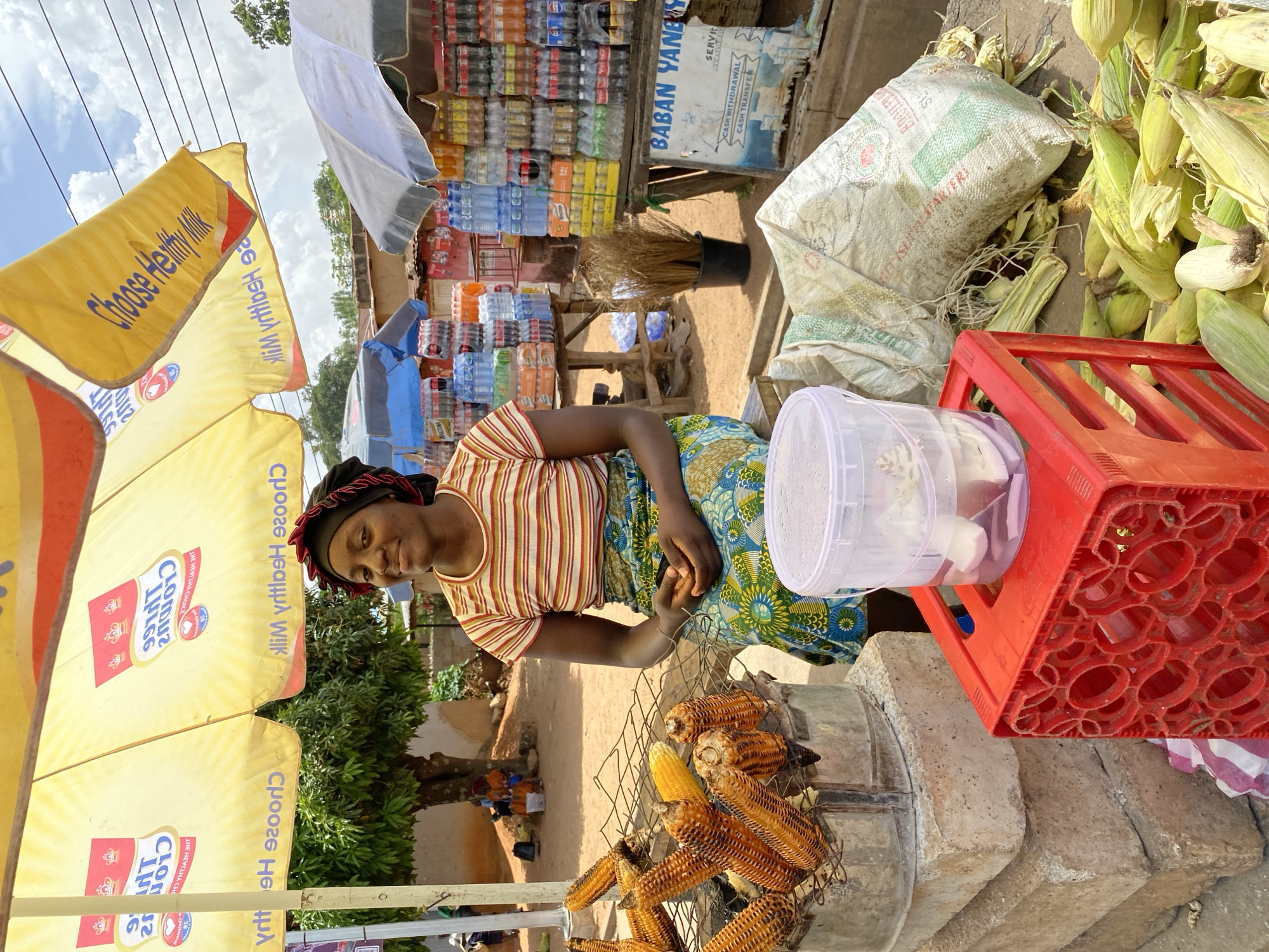 An outdoor scene in Bukuru, Nigeria, captures a woman grilling corn at a roadside stall on a bright, partly cloudy day. The woman, wearing a striped shirt, patterned skirt, and a headwrap, sits beside a charcoal grill, tending to several ears of corn that are cooking. To her right, a red plastic crate holds a clear container with what appear to be peeled garlic cloves.
Behind the woman, to the left, two large yellow umbrellas are prominently displayed, featuring promotional text such as "Choose Healthy Milk" and "CLOUDS." In the background, a busy stall is stacked with numerous bottles of water and various soft drinks. Adjacent to this beverage stall, a blue and white sign advertises "BABAN YANEY" with services like "CASH WITHDRAWAL" and "CASH TRANSFER," indicating a mobile money or financial service agent.
On the right side of the frame, a large pile of fresh corn cobs, some still in their green husks, rests on the unpaved ground next to a partially visible white sack. The woman looks towards the viewer with a slight smile, contributing to the lively atmosphere of the market or roadside vendor setup. The visible text on the umbrellas and the sign provides commercial context to the bustling scene.