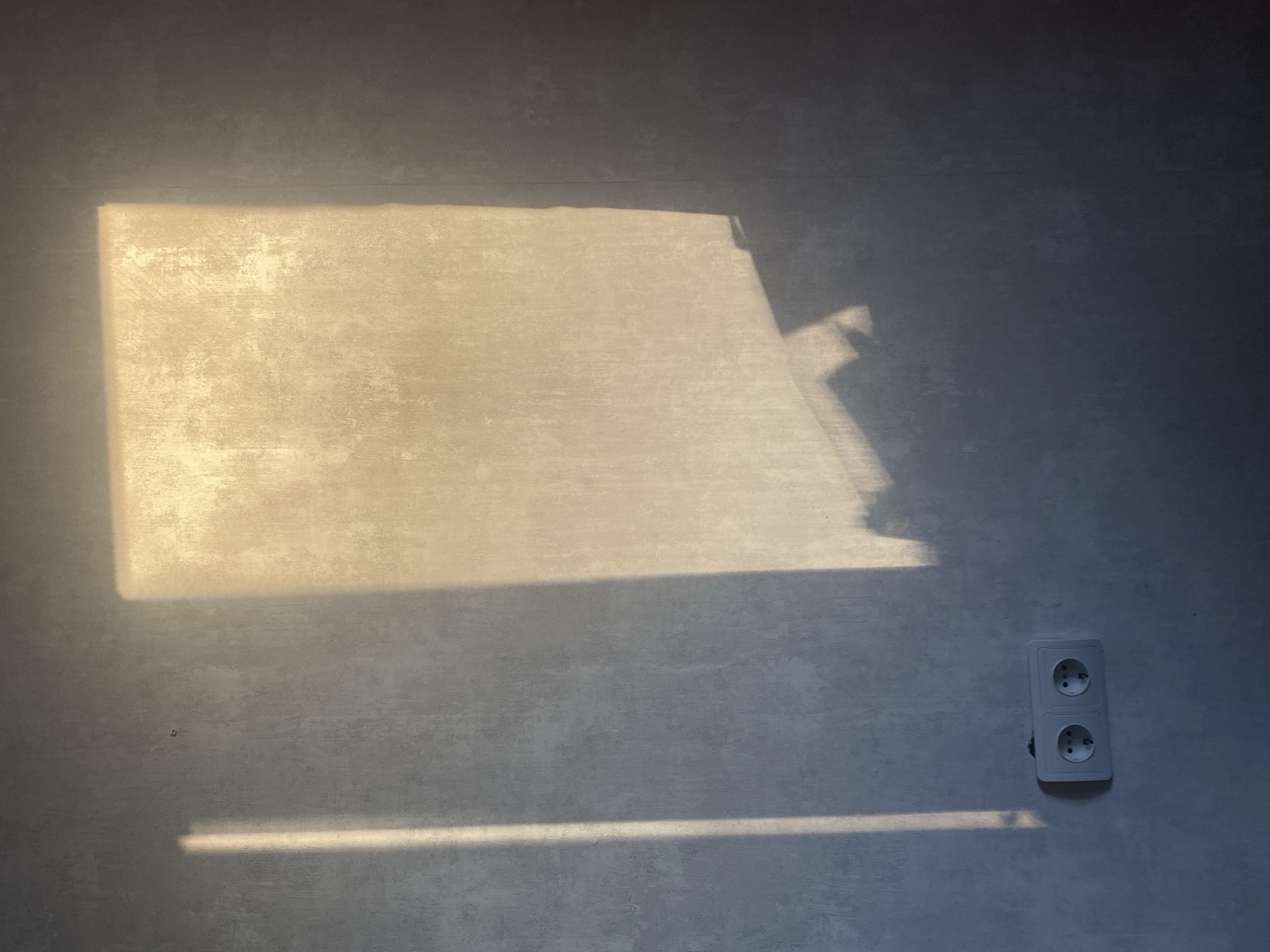 A neutral-toned wall, likely interior, is illuminated by a rectangular patch of sunlight. Below this larger patch, a thin line of light is visible. An electrical outlet is mounted on the lower right of the wall. The image was taken in Chaikovsky, Russia. There are no people or active events visible in the image. The environment is domestic, simple, and focuses on the play of light and shadow on the wall's texture.