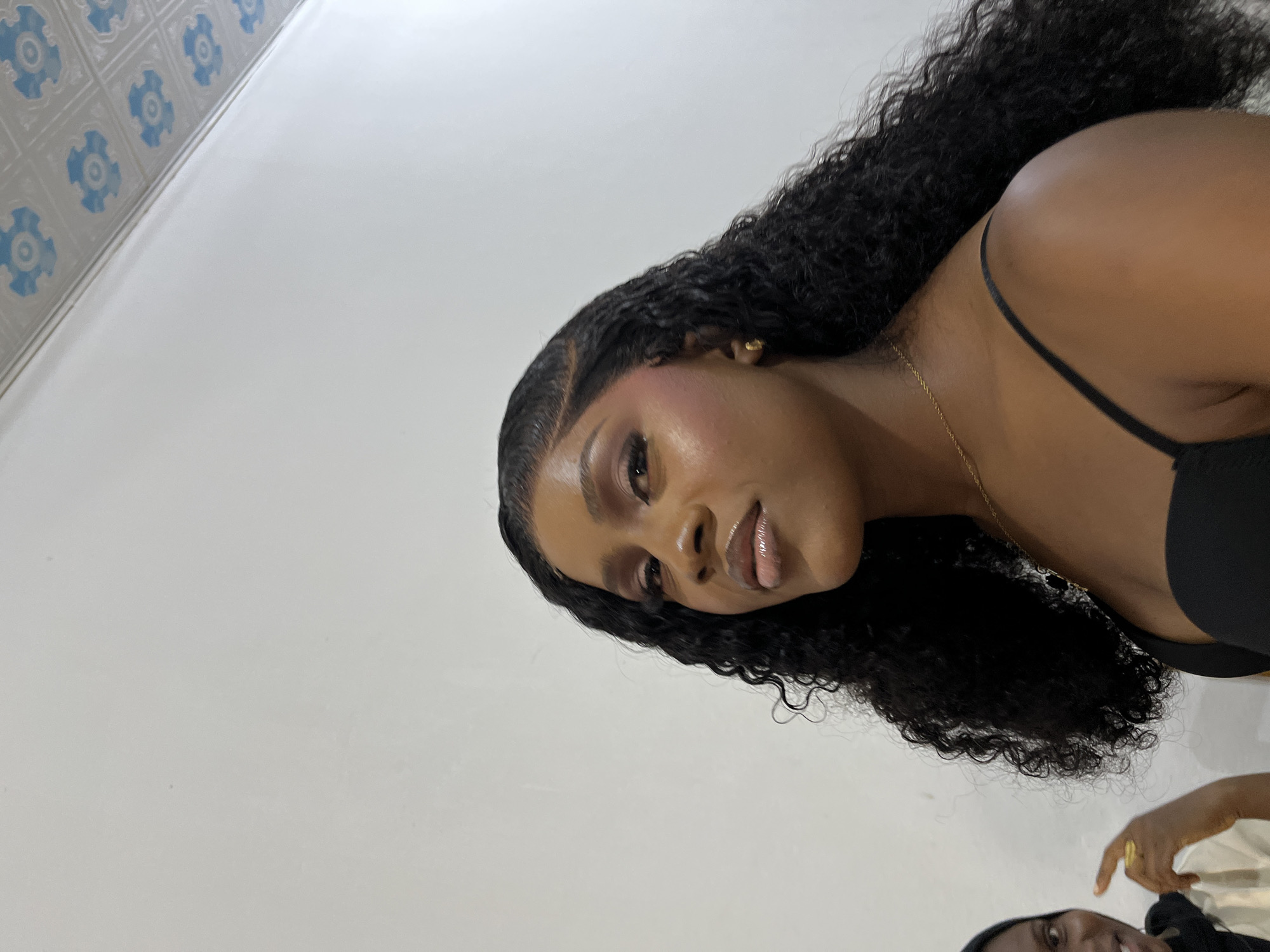 This indoor scene is located in the city of Gura Top, Nigeria.
The primary subject is a woman with a dark complexion, positioned in the center-right of the frame, facing slightly left and upwards. She has long, dark, curly hair, styled with a visible lace front. Her face displays applied makeup, including eyeshadow, blush, and lip gloss. She wears a black spaghetti strap top, a thin gold chain necklace, and a small gold stud earring on her visible right ear. Her expression is neutral.
A second individual, also with a dark complexion, is partially visible in the lower-left corner. This person wears a dark head covering and a light-colored top. Their right hand, adorned with a gold ring, is raised and pointing towards the center of the frame. A portion of their smiling face is visible.
The background consists primarily of a plain, light-colored wall that shows minor surface imperfections and faint specks. In the upper right corner, a section of a tiled ceiling is visible, featuring recurring light blue circular or floral patterns on a lighter background.