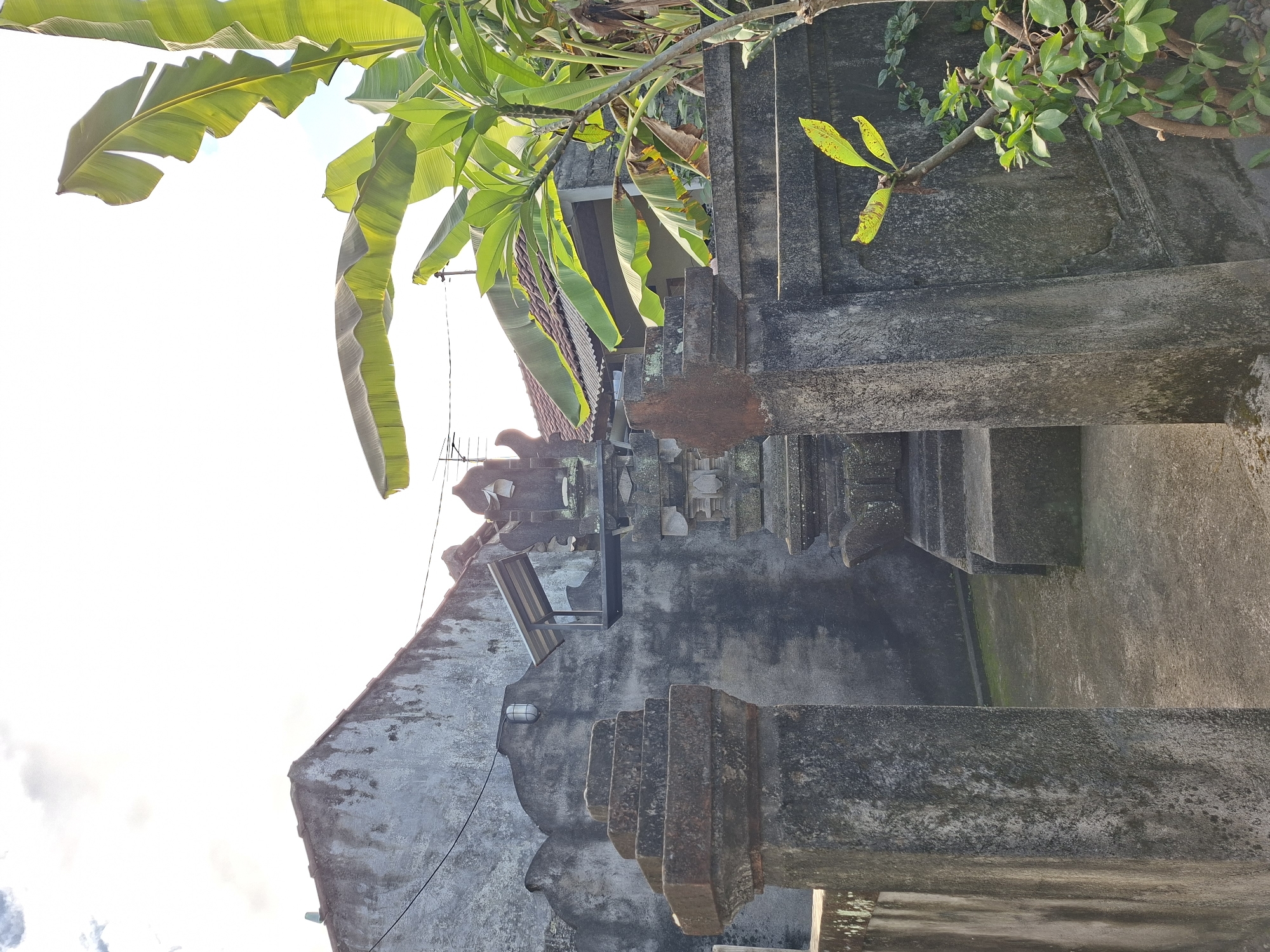 In Blahbatuh, Indonesia, a scene unfolds within a courtyard, possibly a temple or ancestral home, marked by weathered stone structures. The foreground is dominated by two ornate stone pillars, forming an entrance. To the right, a section of a stone wall with decorative molding is visible, framing a small, leafy tree. In the mid-ground, a tiered stone structure, resembling a pedestal or shrine, stands centrally. Behind it, the back wall of a building is textured and aged, with a single light fixture mounted high on its facade. Beyond this, more buildings with tiled roofs are visible, partially obscured by lush, green banana leaves and other foliage. The sky above is bright and hazy, with hints of clouds, suggesting it is daytime.