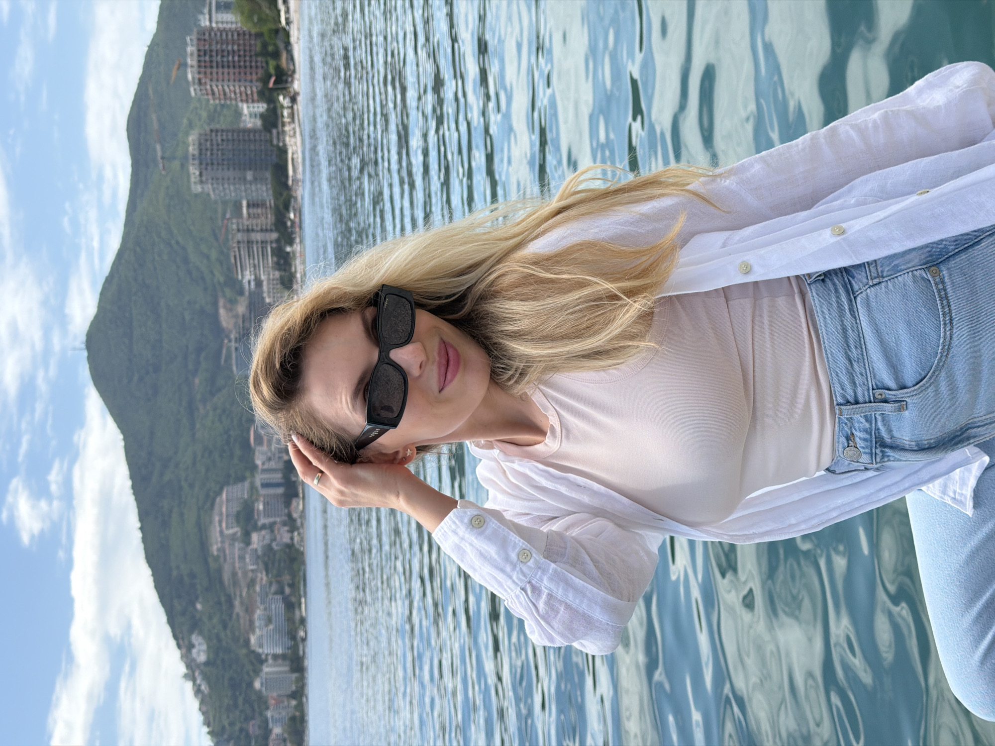 A woman with long blonde hair and sunglasses stands against a backdrop of water and a mountainous urban landscape, appearing to be in Budva, Montenegro. She is dressed in a light-colored shirt worn over a pale pink t-shirt and light-wash denim jeans. Her left hand is raised to her head, fingers touching near her temple, and she is wearing a ring on that hand. The water behind her shows movement with the reflection of the sky and surrounding environment. Behind the water, a steep, green mountain range extends upwards, dotted with buildings along its slope, typical of the Budva area. The sky is visible above the mountains, indicating daylight conditions.