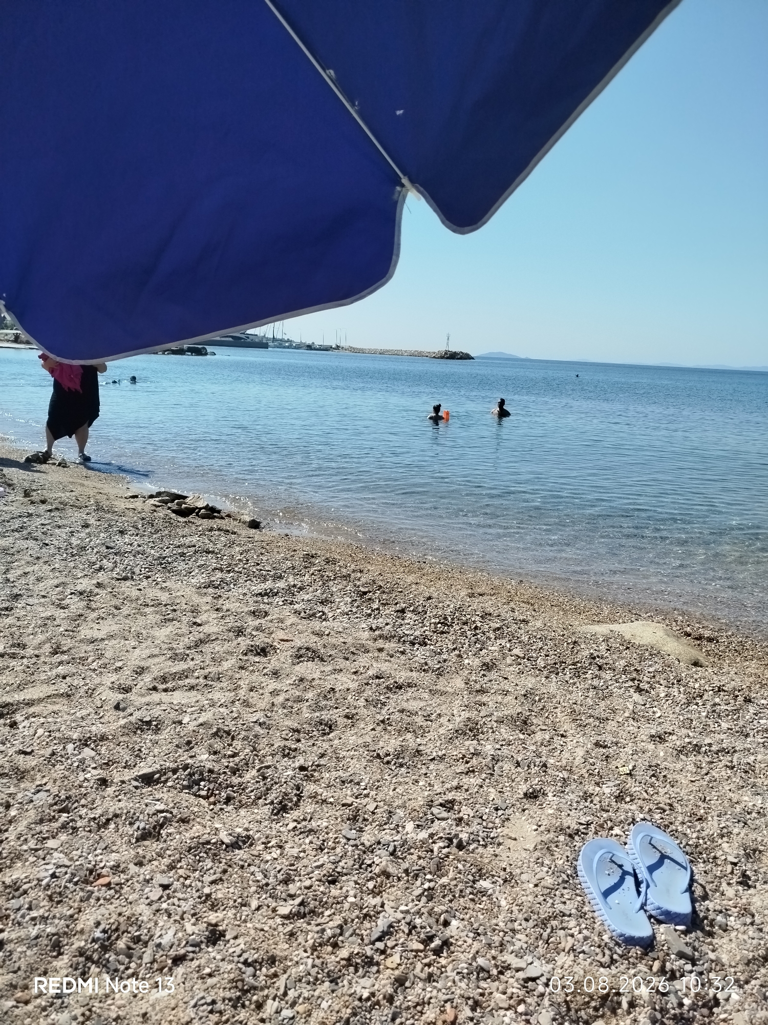 A wide-angle, eye-level shot captures a sun-drenched beach scene in Sithonia Municipal Unit, Greece. In the foreground, a gravelly beach is strewn with small stones and sand. Two light blue flip-flops lie on the beach towards the right side of the frame. A large, partially visible blue umbrella dominates the top portion of the image, casting a shadow over the foreground. In the middle ground, the calm, clear blue water laps gently onto the shore. A person, mostly obscured by the umbrella, stands at the water's edge, wearing a dark skirt and a pink top. Further out in the water, two individuals are swimming. In the distance, a long, rocky pier extends into the sea, with a small lighthouse-like structure at its end. The horizon is clear, with a light blue sky meeting the sea, and faint hills visible on the far horizon. The overall impression is one of a peaceful summer day at the seaside.
