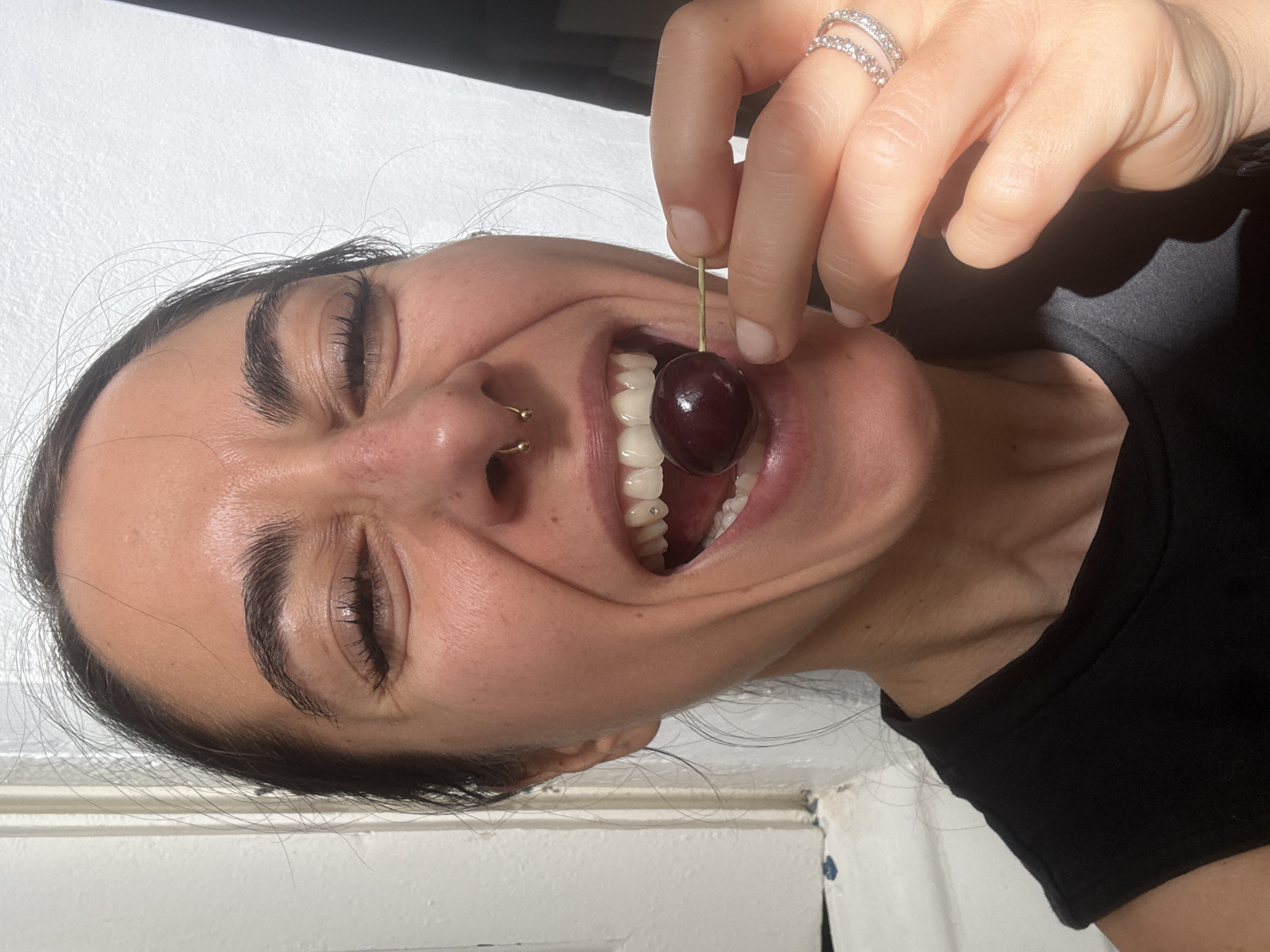 A woman is holding a cherry with a toothpick to her open mouth. She is wearing a black tank top and a ring with diamonds on her right hand. Her hair is dark brown and pulled back. She has a nose ring and a dental gem on one of her teeth. The background is a white wall and a doorway. There is no indication of the city of Barcelona, Spain in the image.