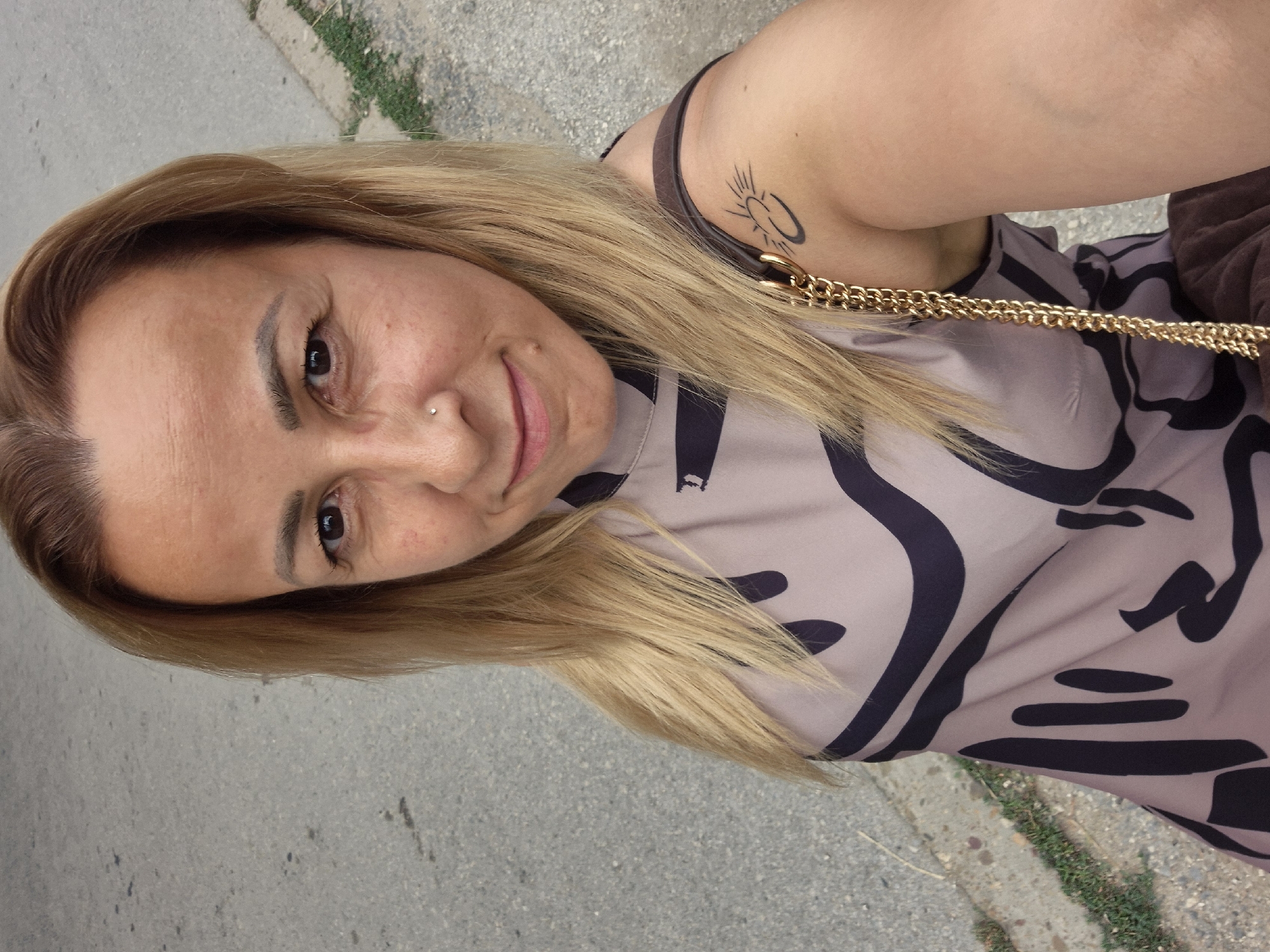 A woman with blonde hair and a nose ring smiles at the camera, taking a selfie outdoors. She wears a light-colored sleeveless dress with a black abstract pattern and a brown bag with a gold chain strap is slung over her right shoulder. A tattoo of a sun-like symbol is visible on her upper right arm. She stands on a paved surface, likely a sidewalk or street, with some grass and a curb visible to her right. The lighting suggests it is daytime.