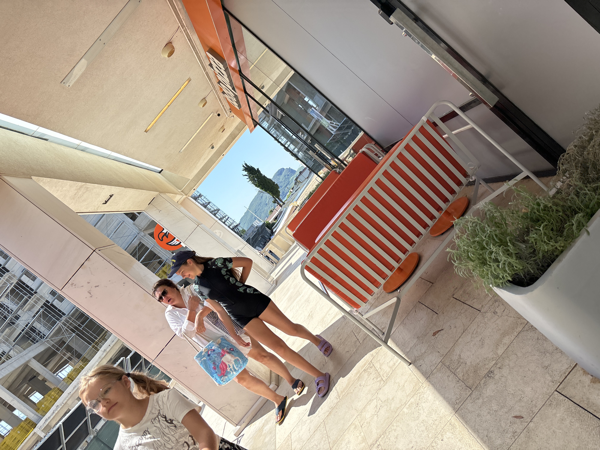 The image depicts three young females walking along a tiled outdoor walkway. The walkway is lined with a modern building featuring large glass windows and an orange awning. To the right, there are bright orange and white striped lounge chairs and a large planter with greenery. In the background, a clear blue sky is visible with mountains and a single evergreen tree.
The person in the foreground, on the left, is a young blonde girl wearing glasses and a white t-shirt. She is looking towards the camera. To her right, two other females are walking. The one in the middle is wearing a white top and sunglasses, holding a colorful tote bag with a unicorn on it. The person on the far right is wearing a dark baseball cap and a black one-piece swimsuit with a leafy pattern. She is looking ahead and appears to be walking with a slight bounce in her step.
The lighting suggests it is daytime, with bright sunlight casting shadows. The scene appears to be a resort or a promenade, given the lounge chairs and the view of the mountains and sea. The overall atmosphere seems relaxed and summery. There is visible text on the orange awning in the background, but it is not clearly legible. The building in the background appears to be under construction, with scaffolding visible.