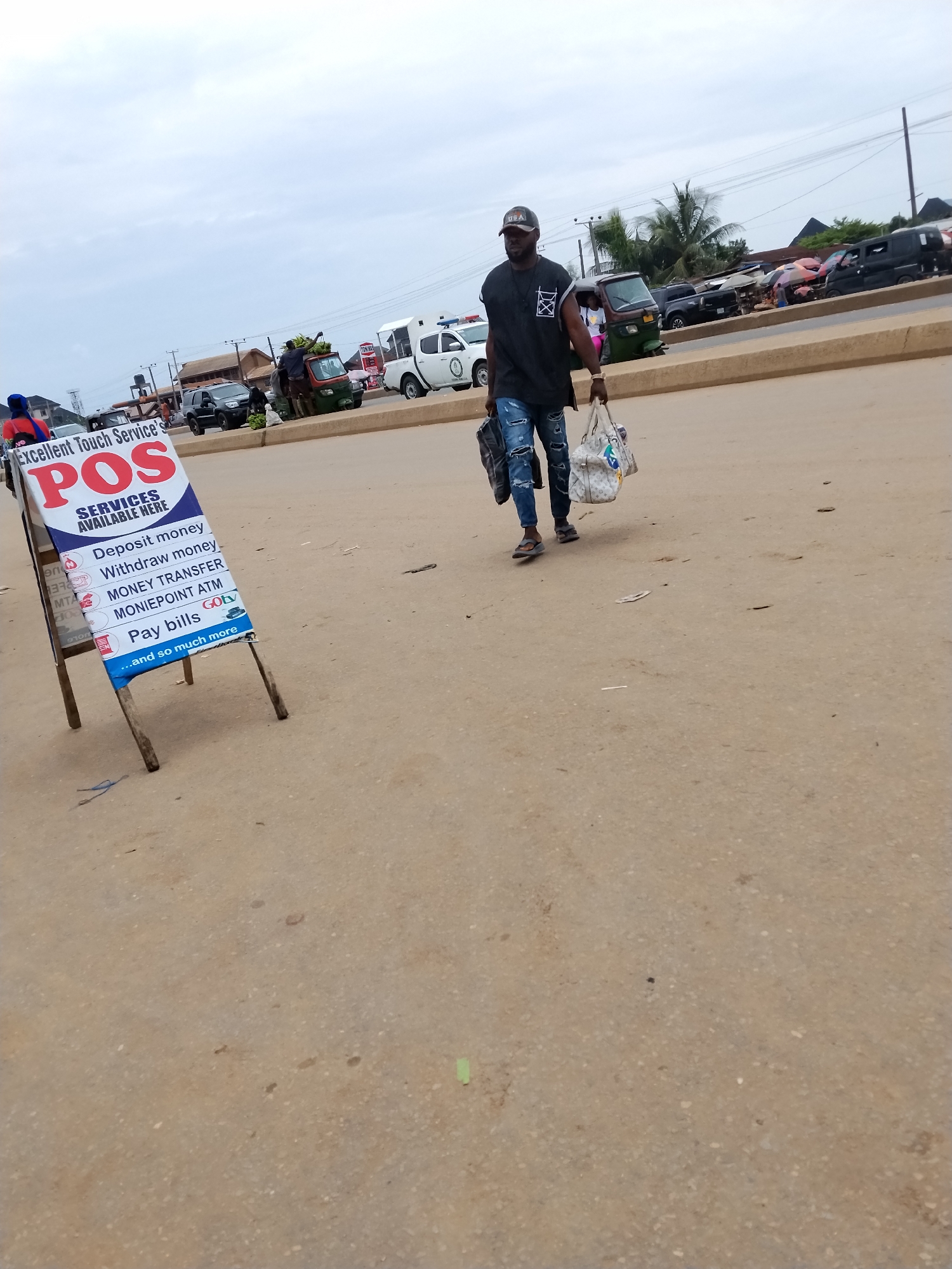 The image captures a street scene in Ura Nta Umuarandu, Nigeria. The location appears to be a roadside, possibly a commercial area, evidenced by vehicles, including trucks and a medical response vehicle, parked along the road. A pedestrian, a man, walks across the road. He carries two bags and wears a cap, a black tank top, and ripped jeans with open-toed shoes. An advertisement sign board stands on the side of the road advertising "Excellent Touch Service POS" services such as money deposit, withdrawal, transfers, ATM, and bill payments. The road surface is worn and somewhat dusty. The sky is overcast.