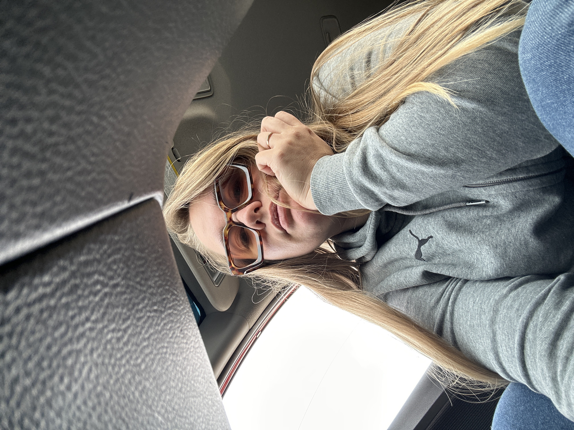 A woman with long blonde hair and sunglasses is seated in a car. She is wearing a grey Puma hoodie and blue jeans. Her hand is near her face, and she is wearing a ring on her finger. The car's interior is visible. There is no information to suggest the location is Ural'skoe, Russia.