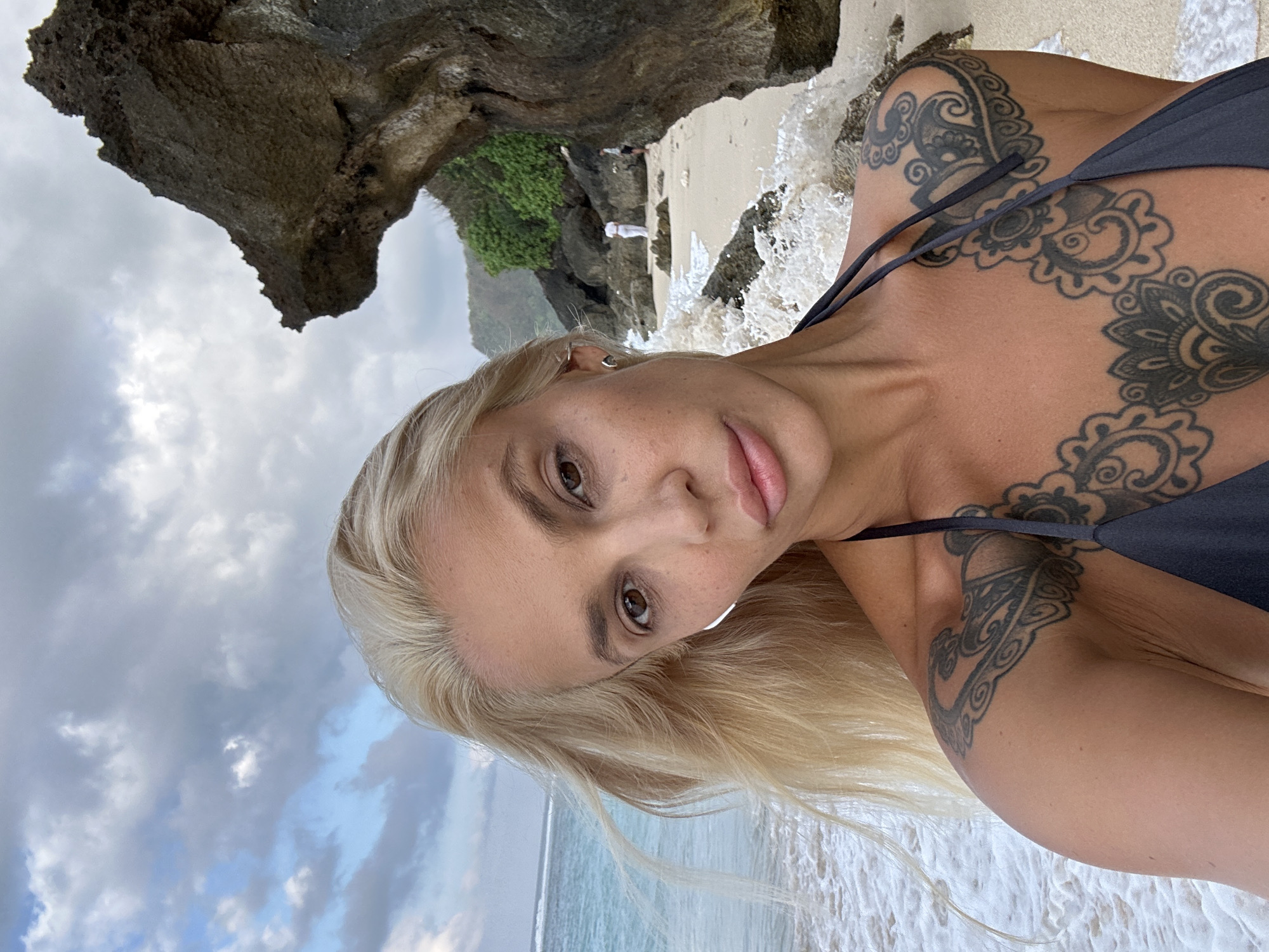 The selfie features a blonde woman with tattoos on her chest and shoulder, wearing a dark bikini top. She is positioned in the foreground with the ocean and a rocky coastline behind her. In the background, two people are visible on the beach near the water. The scene appears to be the beach in Kuta Selatan, Indonesia, characterized by its sandy shore, crashing waves, and lush green vegetation on the cliffs. The sky is cloudy.