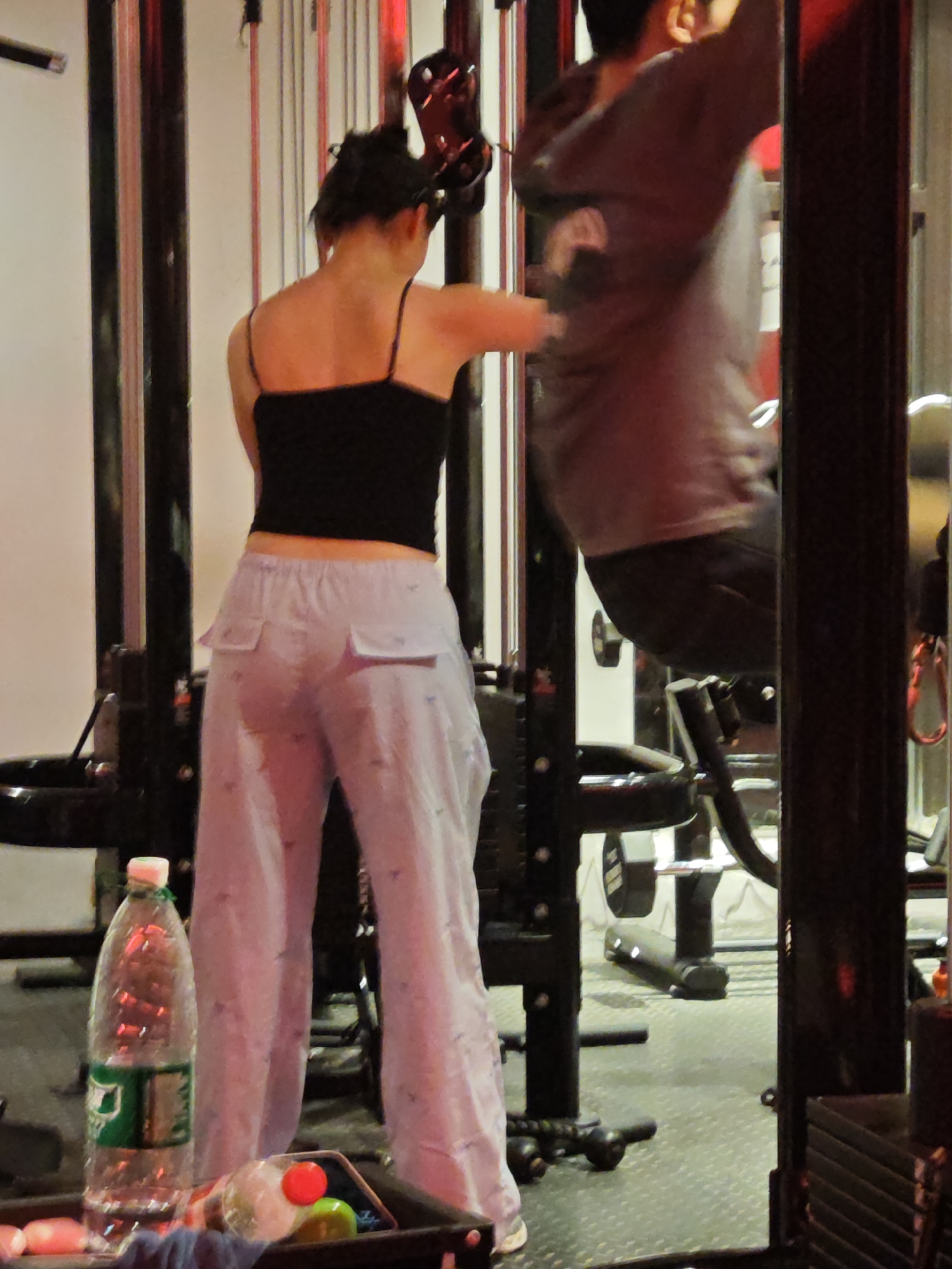 This photograph captures a scene inside a gym or fitness center in the Chongqing High-tech Zone, China, likely during the evening or at night, as indicated by the artificial red and ambient lighting.
Two individuals are present in the frame. In the foreground, facing away from the camera, stands a woman wearing a black spaghetti-strap crop top and loose-fitting, light purple trousers with notable side pockets. Her hair is tied up. She is positioned near a tall, black gym machine, possibly a cable station or pull-up rack.
To the right, partially obscured and blurred by motion, a male figure is actively performing an exercise on a piece of gym equipment. He is wearing a gray long-sleeved shirt or hoodie and dark pants, and his posture suggests he is in the midst of an upper-body workout, such as pull-ups.
In the lower left foreground, a clear plastic bottle of Sprite, clearly identifiable by its green label and white cap with the "Sprite" logo, rests on a dark surface. Beside it, a smaller bottle with a red cap and a green item are also visible. The gym floor is a dark, textured rubber mat. Various other pieces of black gym equipment and weights are scattered throughout the background, completing the fitness environment.