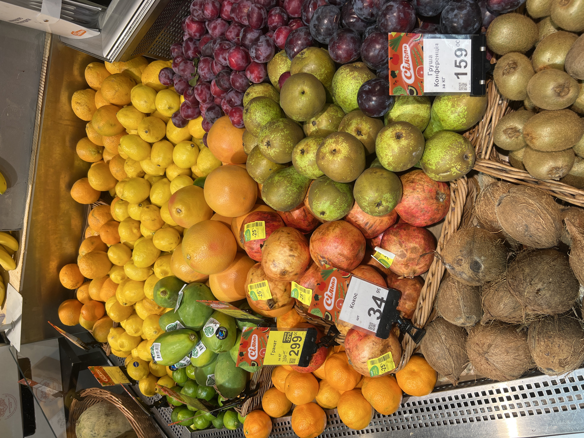 The image shows a produce display in a grocery store, likely in Odesa, Ukraine, given the Cyrillic text on the price tags. The display features a variety of fruits including grapefruits, lemons, limes, papayas, oranges, pomegranates, plums, pears, coconuts, and kiwis. Several price tags are visible, with prices in Ukrainian Hryvnia (UAH). One tag indicates "Гранат" (Pomegranate) at 299.00 UAH, another "Кокос" (Coconut) at 34.99 UAH, and a third "Груша Конференція" (Conference Pear) at 159.00 UAH. The fruits are arranged in woven baskets and metal containers. The background shows more fruit displays and store shelving. There are no people visible in the image.