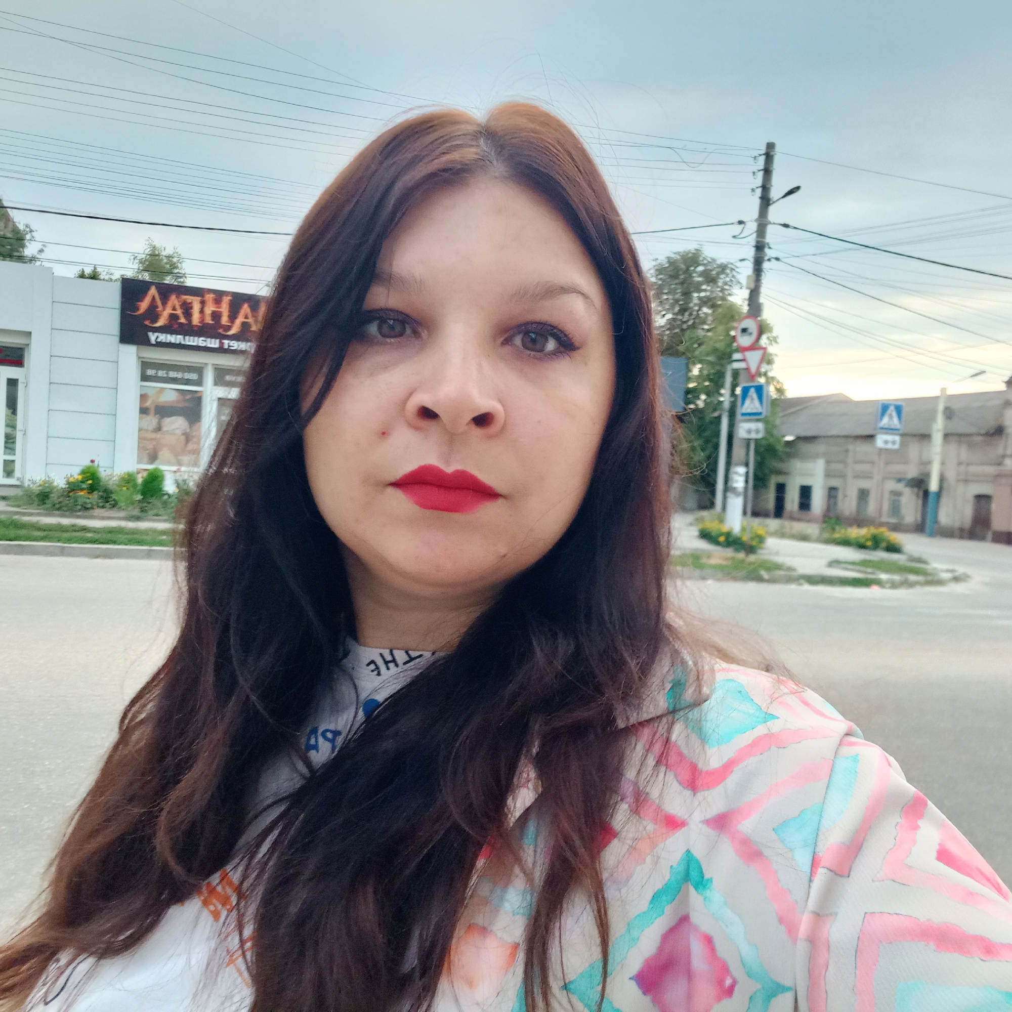 A selfie taken outdoors in Kropyvnytskyi, Ukraine. The subject, a woman with long, dark brown hair and red lipstick, is looking at the camera. She is wearing a light-colored shirt with a geometric pattern. In the background, there is a building with a sign that appears to say "AATHA" in large, stylized letters. The building also has windows displaying what looks like baked goods. Further in the background, there are street signs, a utility pole with numerous wires, trees, and a few buildings with a muted color palette, suggesting an urban setting. The sky is overcast, indicating a cloudy day.