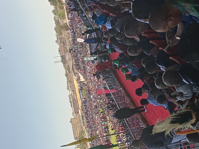 This image captures a large outdoor public gathering or rally, likely held in Siaya, Kenya, viewed from an elevated perspective. The scene is filled with a vast crowd of people extending into the distance under a clear, bright sky, indicating daytime with sunny weather.
In the foreground, individuals are visible from above, many seated in red stadium-style seats. Their heads and backs are mostly visible, with some appearing to look forward or downwards, engrossed in the event. A broad red carpet or covered area forms a prominent path or stage extending from the immediate foreground into the mid-ground.
The mid-ground is densely packed with attendees, forming a sea of people. Interspersed within the crowd are several banners and flags. A red banner with white text is clearly visible, displaying "LUO" and the year "2013". Other banners, including one with green and white colors, are also present but their text is illegible. Two tall, slender poles or masts rise above the crowd in the mid-distance.
The background reveals a flat, open landscape with some distant buildings, consistent with an urban or semi-urban environment. A faint, thin line, possibly from a kite or a distant aerial object, is visible against the clear sky in the upper left. The overall impression is one of a significant, well-attended public assembly.