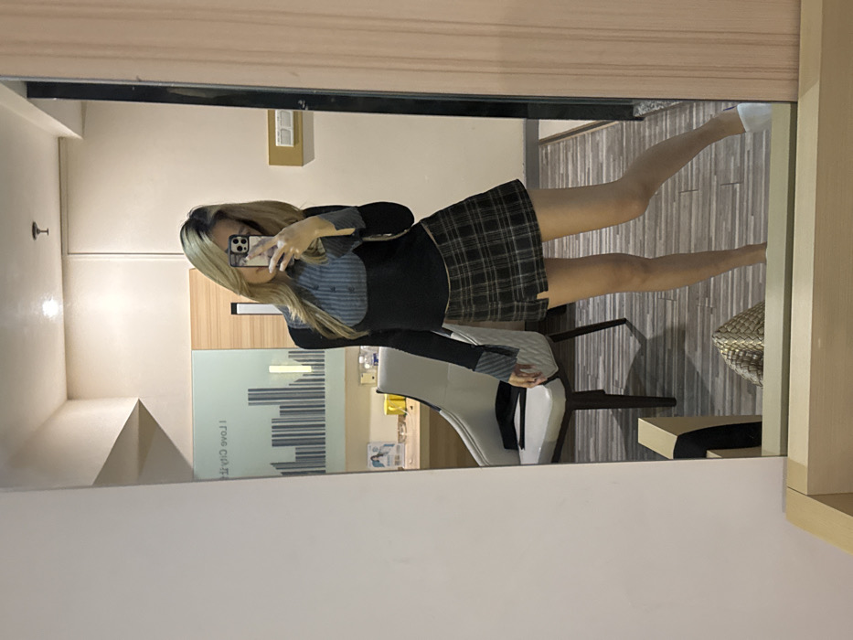 A female individual is depicted in a mirror selfie taken indoors, within a room likely situated in Yuzhong District, China. The individual, possessing long, light-colored hair, stands frontally to the mirror, holding a dark-colored smartphone to capture the image, which partially obscures her face. She is dressed in a long-sleeved top featuring a black torso section with an integrated grey and white pinstriped collar, placket, and matching cuffs. She wears a dark plaid mini-skirt with a front slit. A white slipper is visible on her right foot. The reflected background reveals a light-colored armchair with a dark strap draped over its seat, positioned to the left. Behind the armchair, a desk or counter surface holds several indistinct items. Above this, a wall panel displays a grey and white stylized cityscape motif, with the partially visible reflective text "I love City" above it. A woven, golden-colored handbag rests on the horizontally striped, light-colored wooden floor to the right of the armchair. A white wall-mounted light switch or outlet panel is visible on the wall to the right of the mirror's frame.