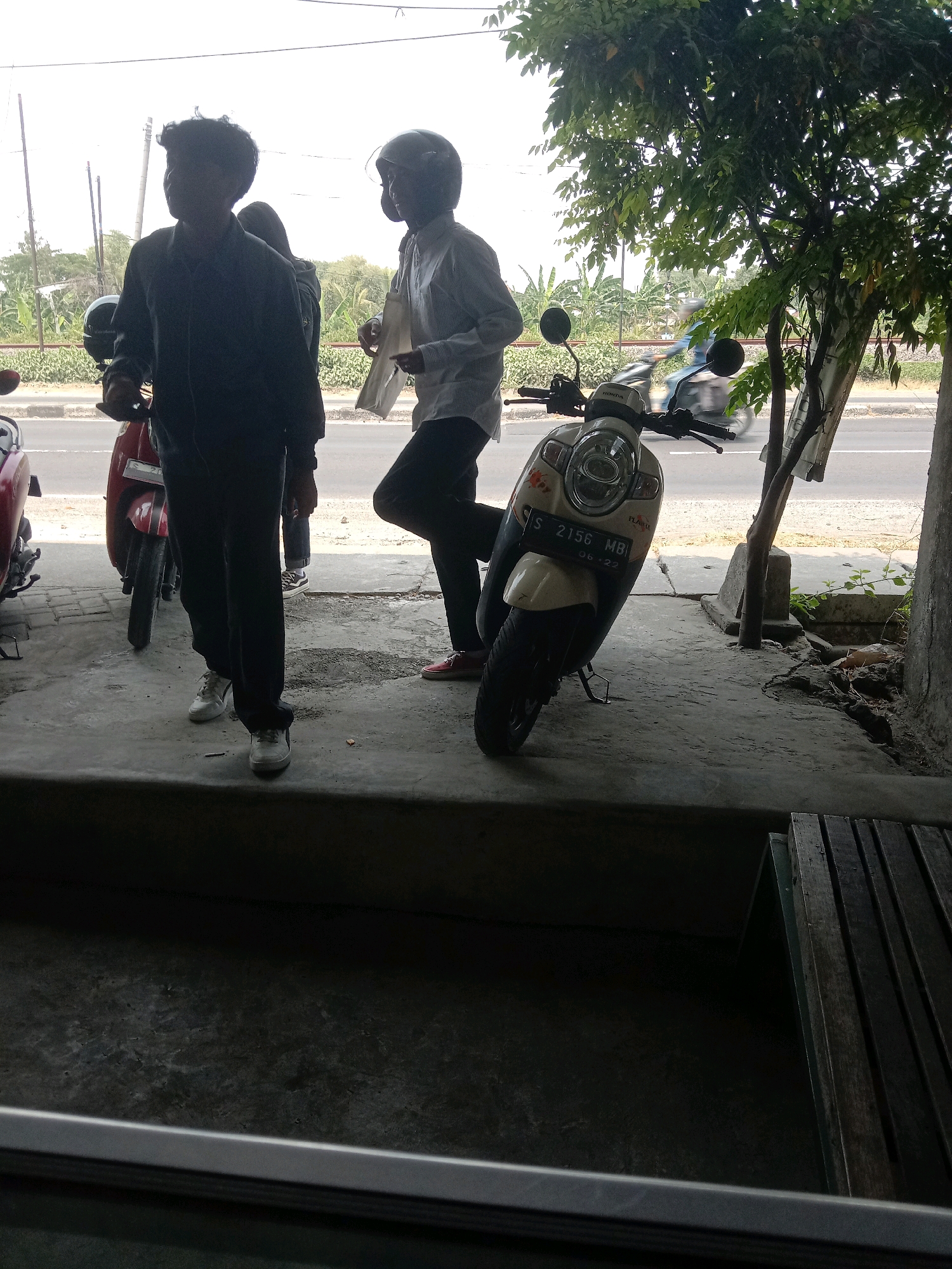 In Plosowahyu, Indonesia, two individuals are standing near a street with motorbikes. The person on the left, silhouetted against a bright sky, appears to be wearing dark clothing and white sneakers. To their right, another person in a light-colored shirt and dark pants, also wearing sneakers, stands with a helmet on their head. A white and beige motorbike is parked in the foreground, with a license plate that reads "S 2156 MBI 06-22". A red motorbike is partially visible to the left of the frame. The environment suggests a roadside setting with a concrete surface and some vegetation. The sky is overcast, and the lighting is diffuse.