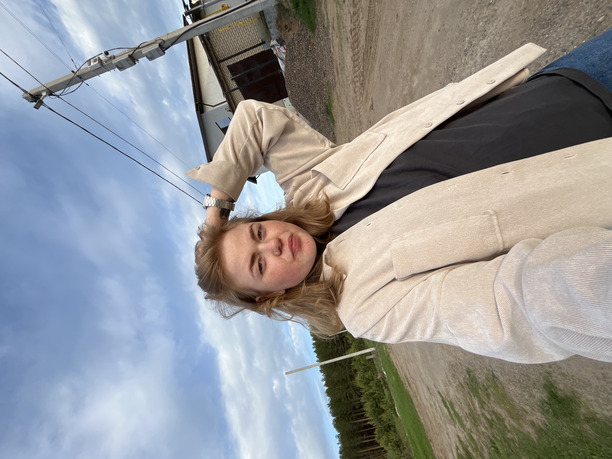 A young woman with fair skin and freckles is posing outdoors in Chaikovsky, Russia. She wears a black shirt and a tan, corduroy jacket. She also has a silver watch. She has her hand resting behind her head. Overhead, a utility pole and building can be seen. The ground appears to be a dirt road. In the distance, a line of trees is visible. The sky is overcast.