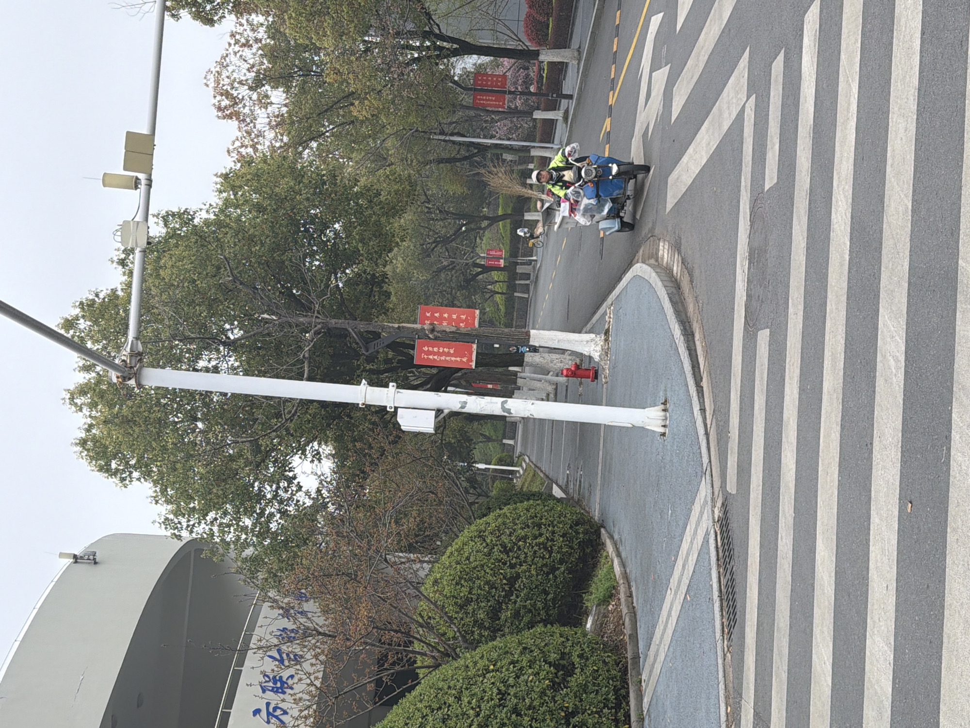 The image shows a street scene in what appears to be a developed area, potentially Jiangning District, China, judging from the Chinese text visible on signs. A man is riding a small vehicle, possibly an electric tricycle, loaded with various items including what looks like a broom. The vehicle is traveling on a street that has a crosswalk marked with white stripes. The road also has yellow lane markings. Along the side of the road, there are trees, bushes, and red banners or signs attached to poles. A tall white pole with various pieces of electronic equipment attached runs up the left side of the image. Part of a building is visible in the lower left corner. The weather appears overcast.