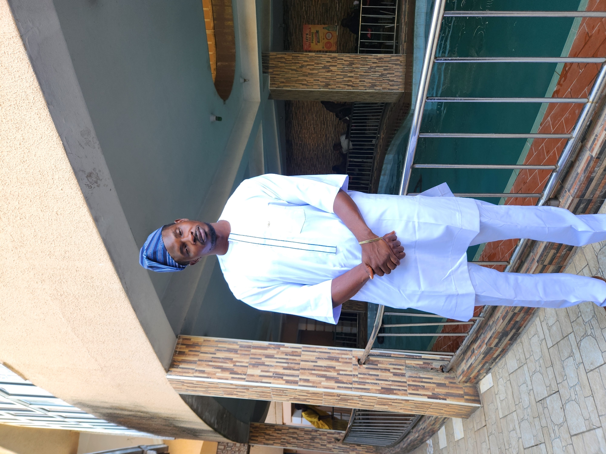 A man wearing a white traditional Nigerian outfit and a blue and white striped cap stands on a tiled floor with a low wall behind him. The wall has a railing and a section with brown and tan tiles. Behind the wall, there is a view of an interior with a sign. The background also shows architectural elements of a building, including pillars and tiled walls. The location appears to be a building exterior, possibly in Unknown, Unknown.