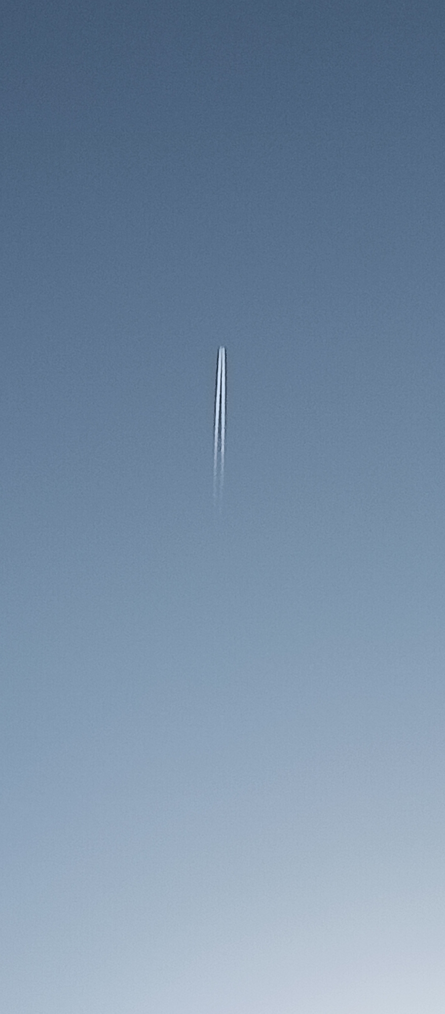 A contrail, likely from an aircraft, is present in a clear, light blue sky. The contrail is vertically oriented and positioned approximately in the center of the frame. No specific details are discernible about the location, city, or time. No other objects or people are visible.