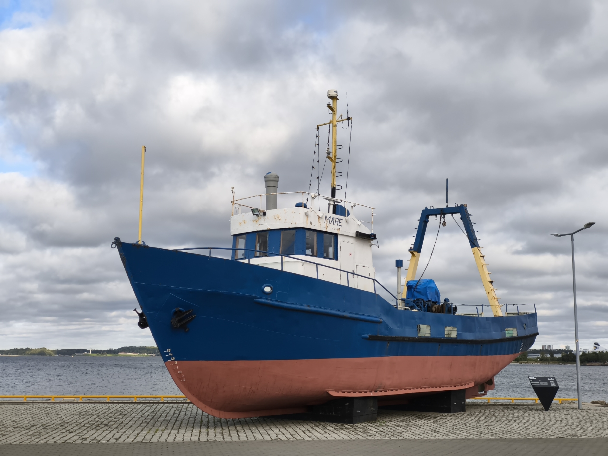 A blue and red fishing trawler named "MARE" rests on a paved embankment near the water in Tallinn, Estonia. The sky is overcast with heavy gray clouds. The trawler is positioned on its hull, supported by black blocks. In the background, across the calm water, a tree-lined shore and distant buildings are visible. A lamppost stands to the right of the vessel, and a black triangular information sign is placed in front of it. The boat's deck features a prominent yellow crane structure.