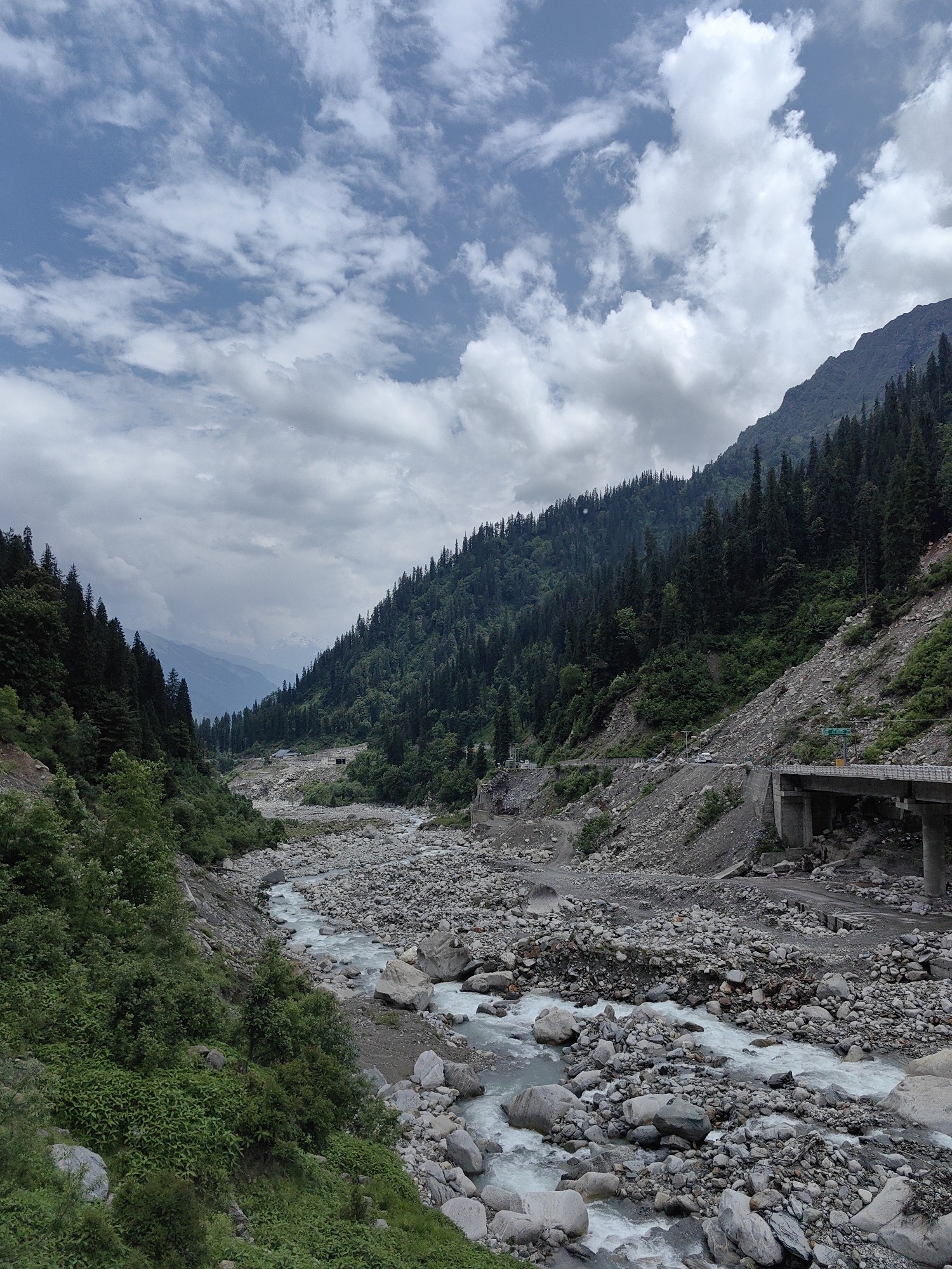 The image shows a mountain valley in Dhundi, India, with a river flowing through it. The valley is covered with trees and rocks. The sky is cloudy. There is a bridge and a road in the image. The river is surrounded by rocks and boulders. The trees are green and dense. The sky is blue with white clouds. There are some buildings in the distance.