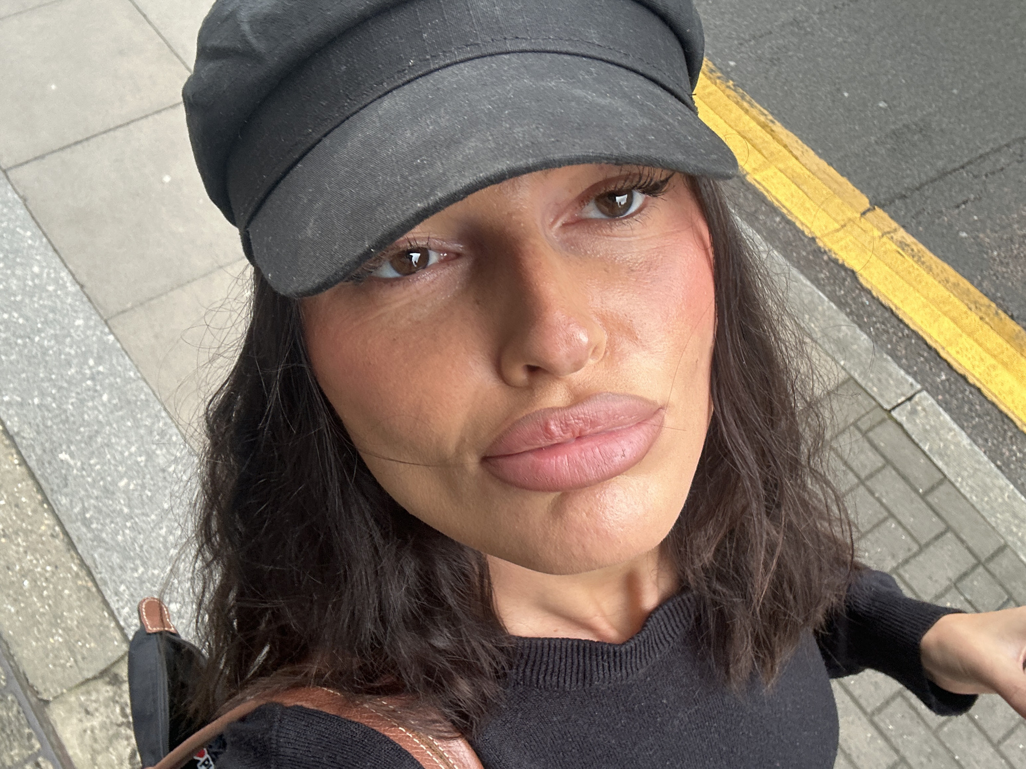 A portrait of a woman is taken from a low angle. She is wearing a black knit sweater, a black newsboy cap, and black socks. Her dark hair is visible. She is outdoors, with a sidewalk and a yellow painted line on asphalt visible behind her. The overall lighting suggests it is daytime. The image is oriented vertically, with the woman's head towards the top of the frame.