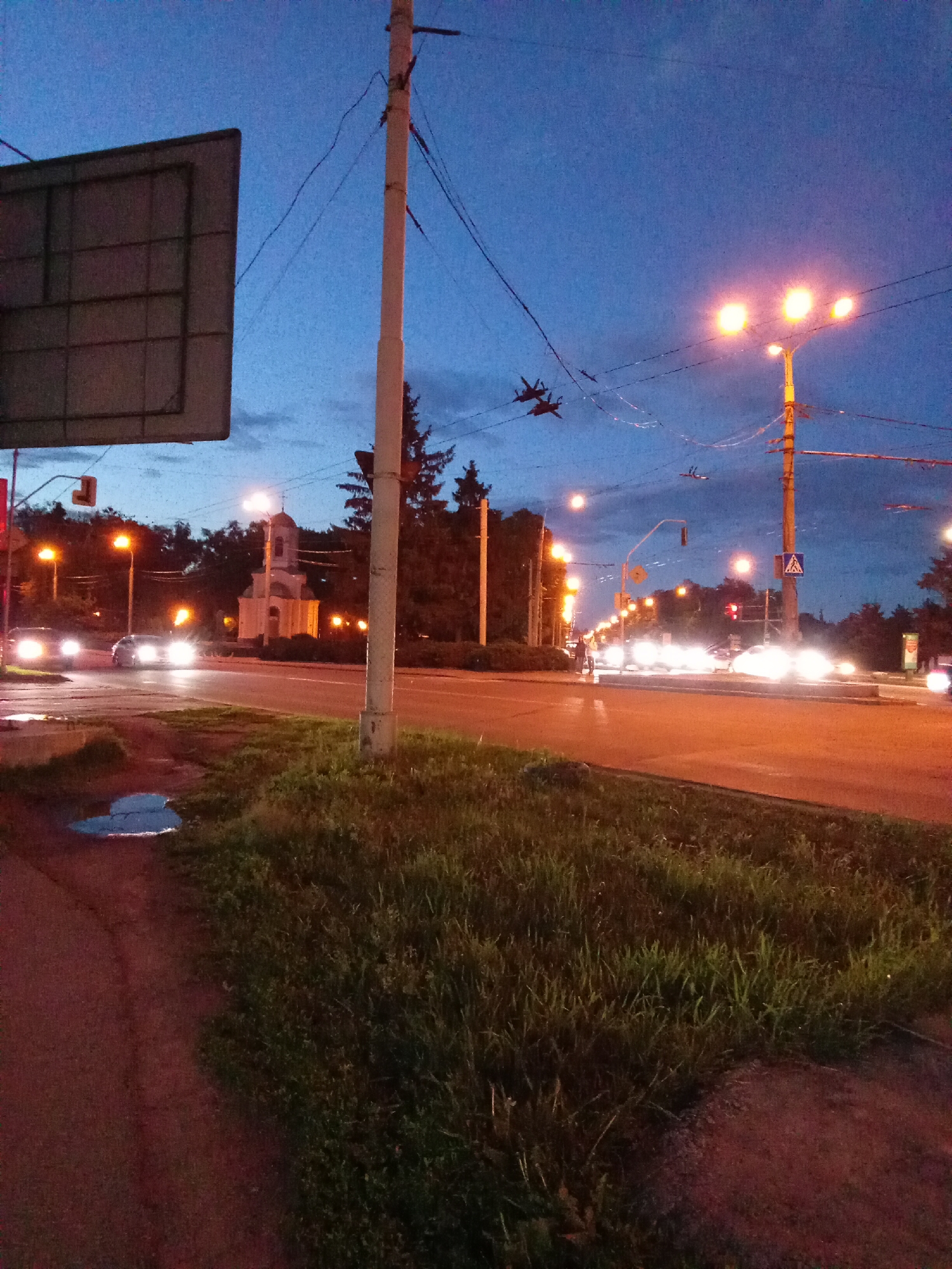 The image depicts an urban street scene at dusk in Poltava, Ukraine. A multi-lane road illuminated by streetlights stretches into the distance. Vehicles are visible on the road with their headlights creating blurred streaks. On the left side of the road is a church, illuminated by artificial lights. Overhead, two military jets fly across the sky against the twilight. A grassy area with overgrown vegetation is in the foreground, with a small puddle on the sidewalk. A utility pole and signage are on the left side of the frame.