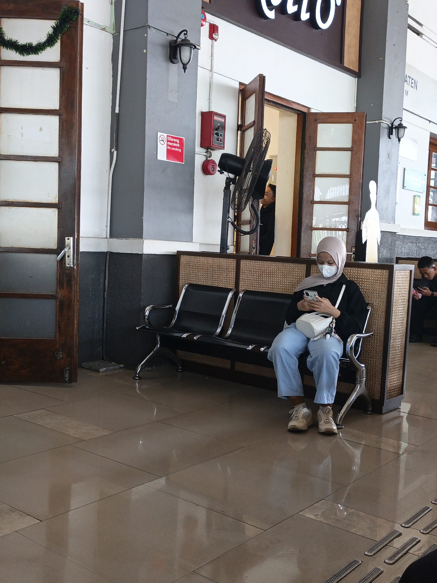 The image depicts a waiting area inside a building, possibly a train station, in Klaten, Indonesia. In the foreground, a young woman wearing a hijab and a face mask sits on a black bench. She is dressed in a black top and light blue pants, with white sneakers. She is holding and looking at a smartphone. Beside her is a white handbag. The bench is part of a seating arrangement with a woven partition behind it.
To the left of the woman, a wooden door with glass panels is ajar, revealing a hallway beyond. Above the door, a garland of dark green tinsel hangs. A gray column is visible to the left of the column, with a "No Smoking" sign in red and white on it. A black metal standing fan is positioned in front of an open doorway further back, where another person is partially visible.
To the right, another person, a young man, sits on a similar bench, also looking at a smartphone. The floor is tiled with large, light brown squares that reflect the light. The overall environment suggests an indoor public space, likely a transportation hub. The architectural style and the presence of the "No Smoking" sign in Indonesian confirm the location as Indonesia, with Klaten being a specified city.