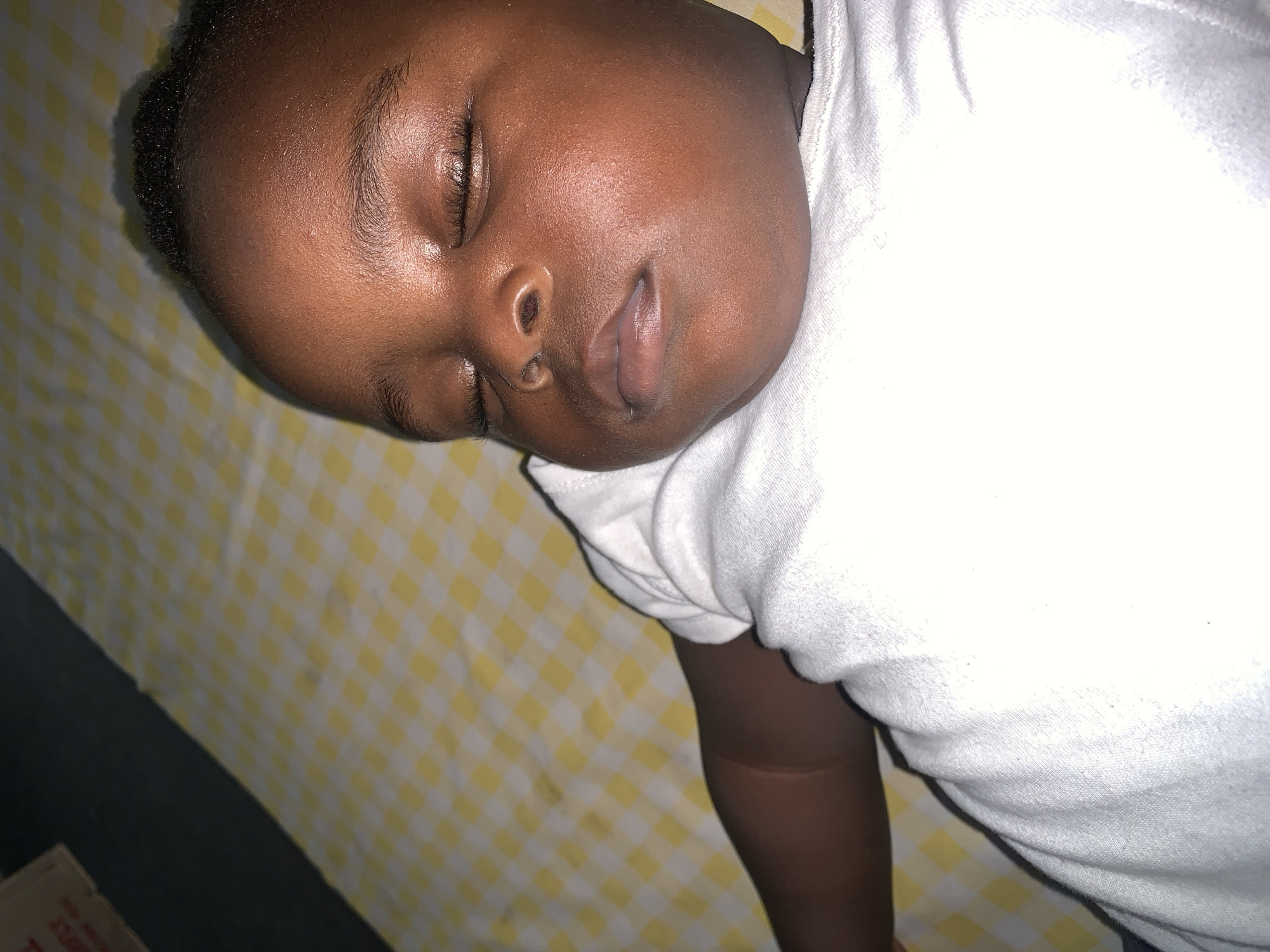 A close-up, overhead shot captures a Black baby sleeping peacefully on a yellow and white checkered surface. The baby is wearing a white t-shirt. The lighting is warm, and the background is slightly blurred, focusing attention on the sleeping infant. The scene appears to be indoors, in an unspecified urban environment.