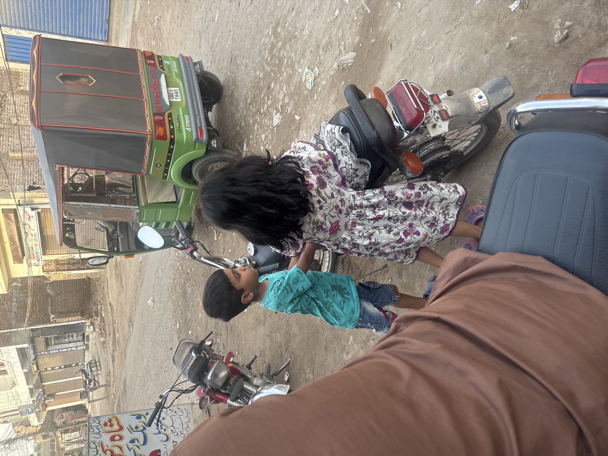 The image depicts an outdoor street scene in a city of Unknown, Unknown, characterized by unpaved ground and buildings. In the foreground, the lower portion of a person wearing a brown garment is visible on the bottom left, positioned as if seated on a vehicle. Directly in front, a young boy, approximately 4-6 years old, with dark hair, is standing to the left of a silver and red motorcycle. He is wearing a teal short-sleeved t-shirt with a dinosaur print and denim shorts. His hands are near the motorcycle's handlebars, and he is looking towards the right. To the right of the motorcycle, a young girl, also approximately 4-6 years old, with long dark hair, stands with her back mostly to the camera. She is wearing a white dress with a purple and green floral pattern and pink sandals. Her hands are near the motorcycle.
The central motorcycle is silver and red with a black seat, and "KHAN" is visible on its rear fender. Another red motorcycle is parked further back on the left. On the right side of the background, a green and black three-wheeled auto-rickshaw, branded "NEW ASIA" and marked "4 STROKE," is parked. Its license plate reads "GBG 7645." The ground is uneven, composed of dirt and gravel, with scattered debris. In the background, a row of low-rise buildings with brick and painted facades line the street, many featuring closed metal shutters over storefronts. A white sign with red and black Urdu script, including "شاہ رنگ ورکس," is partially visible on the left side of the scene. The lighting indicates daytime.
