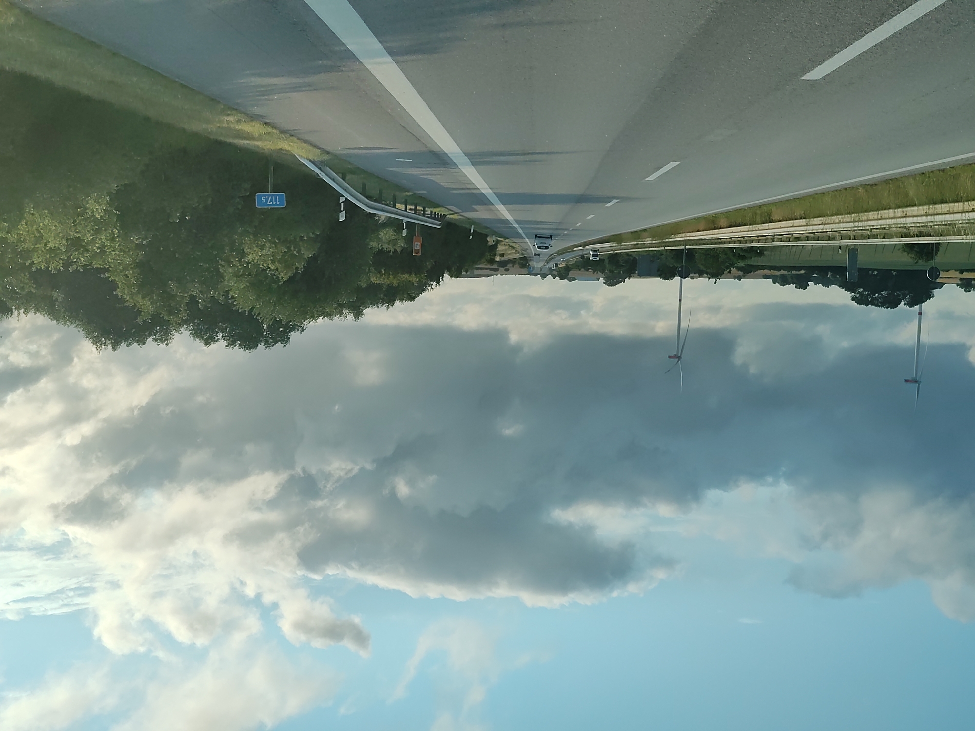 The image shows a highway in Gremersdorf, Germany. The highway is paved and has two lanes in each direction. There are guardrails on both sides of the road. On the left side of the road, there is a field with some trees and two wind turbines in the distance. On the right side of the road, there is a dense forest. The sky is cloudy and overcast, with some patches of blue sky visible. There is a sign on the right side of the road that reads "117.5". There are no people or vehicles visible in the image, except for a white car in the distance on the highway. The image appears to have been taken from a moving vehicle.