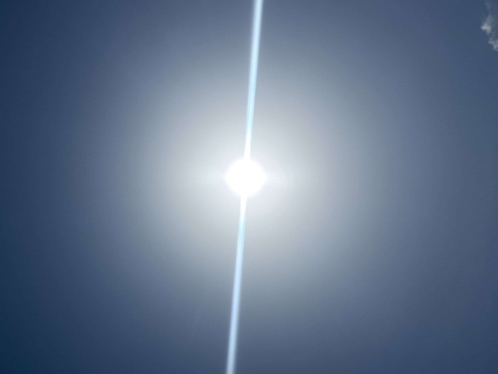 The image presents a bright, overexposed sun in a clear, blue-gray sky, likely captured in Ihiagwa, Nigeria, during the daytime. The sun appears as an intensely bright, pure white circle at the center of the frame. A prominent lens flare manifests as a strong, luminous vertical beam of light passing directly through the sun, extending almost the entire height of the image.
The sky exhibits a gradient, transitioning from a very light, almost white-blue hue immediately surrounding the sun and along the lens flare, to a darker, deep blue-gray in the upper and lower corners and edges of the frame. The overall atmosphere appears clear, with minimal cloud cover; a very small, wispy cloud fragment can be faintly discerned in the upper right corner. The harsh brightness of the sun suggests it is likely midday or early afternoon. There are no other discernible objects, people, or activities visible, focusing solely on the sun and the sky. No text is visible in the media file.