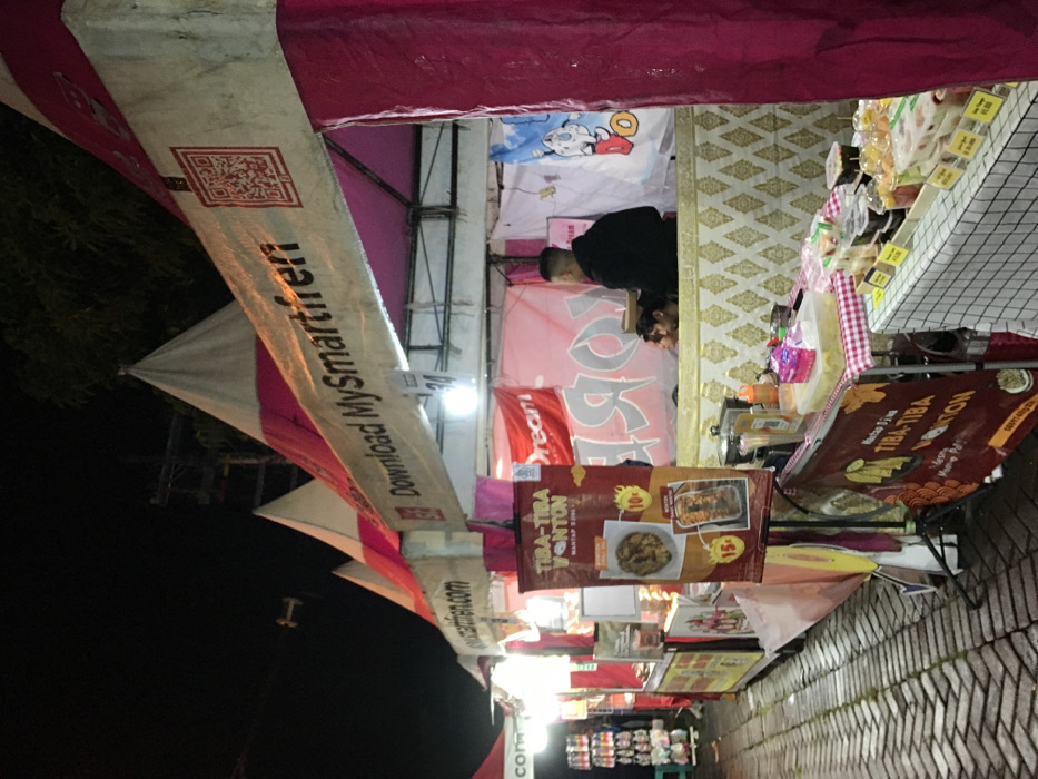 A food stall is visible, with a pink and white awning and banners advertising “MySmartfren” and “TIBA-TIBA WONTON." The wonton sign highlights a price of 10 or 15, presumably in local currency. A person in a black hoodie is seen inside the stall, possibly serving customers. Various food products are displayed on the table. A tiled surface suggests this is a sidewalk location. The environment is dimly lit, indicating it is likely evening or night.