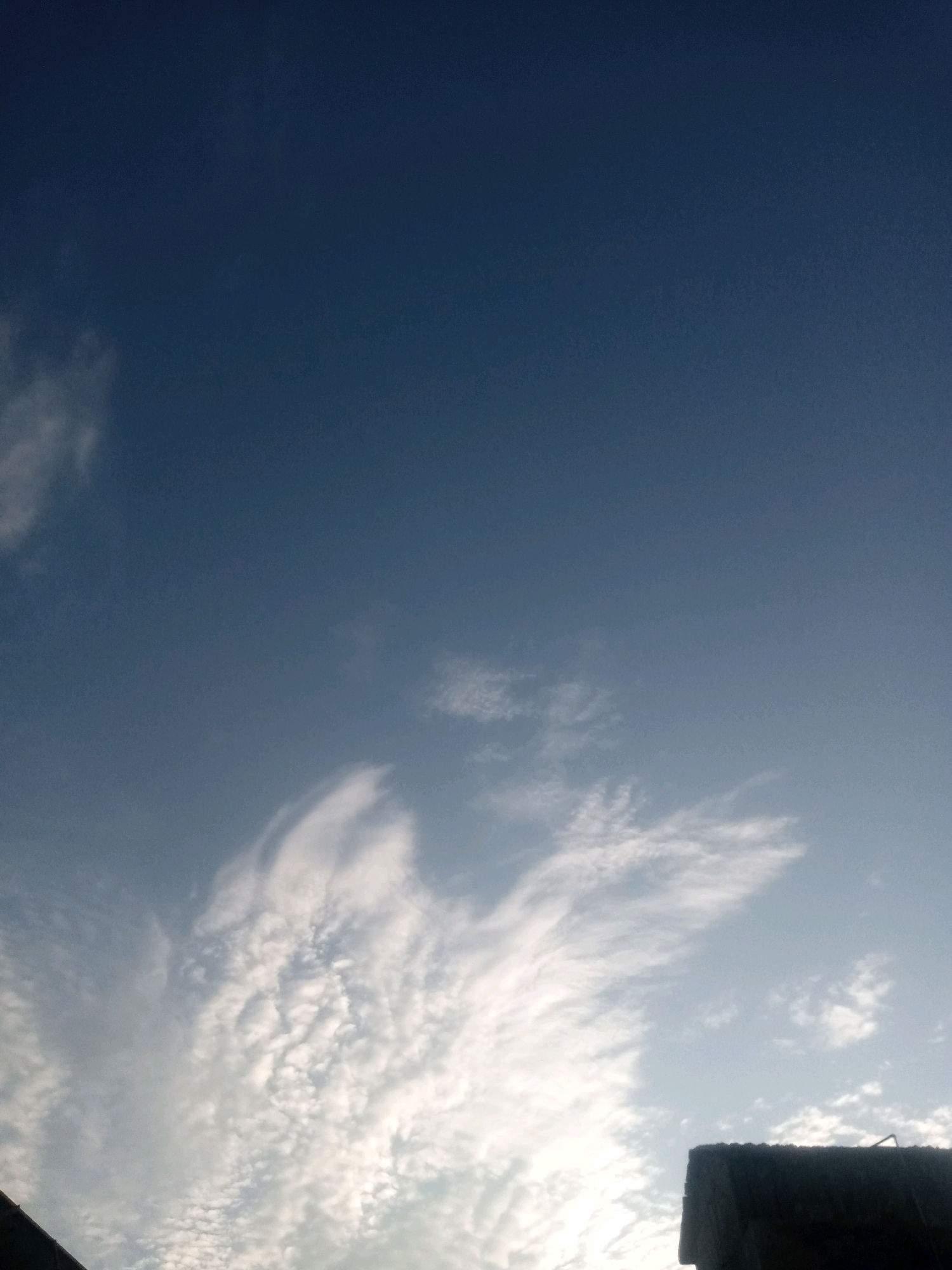 The image captures a sky scene, likely in Tando Jam, Pakistan, with a clear blue expanse dominating the upper portion. Scattered white clouds, some wispy and thin, others more textured and clumpy, populate the lower half of the frame. The sunlight appears to be diffused, suggesting it could be early morning or late afternoon. In the bottom right corner, a dark, block-like structure, possibly part of a building or rooftop, is visible, casting a shadow. The lower left corner shows a sliver of a dark, diagonal edge, possibly another structure or the edge of the frame. No people or animals are present in the image. The overall impression is one of a serene sky with hints of urban elements at the periphery.
