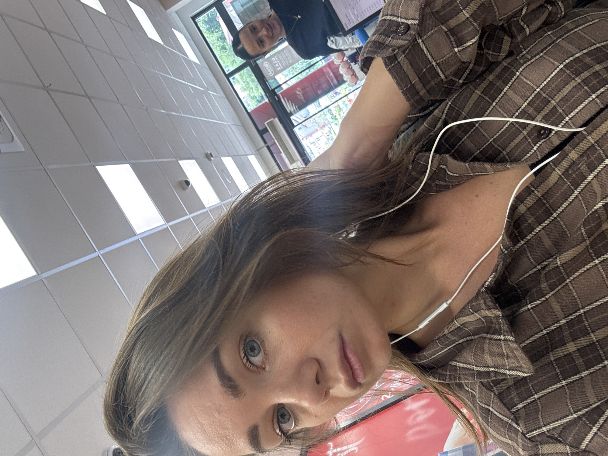 A woman is posing for a selfie in what appears to be a retail environment. She has light brown hair, fair skin, and blue eyes, wearing a brown plaid shirt with white headphones around her neck. In the background, a man with short dark hair, wearing a black shirt, is visible near a window, possibly interacting with a counter or register area. The environment includes a ceiling with fluorescent lights and what looks like a promotional display behind the woman. The location is likely a retail store in Budva, Montenegro, given the red signage with Cyrillic lettering partially visible.