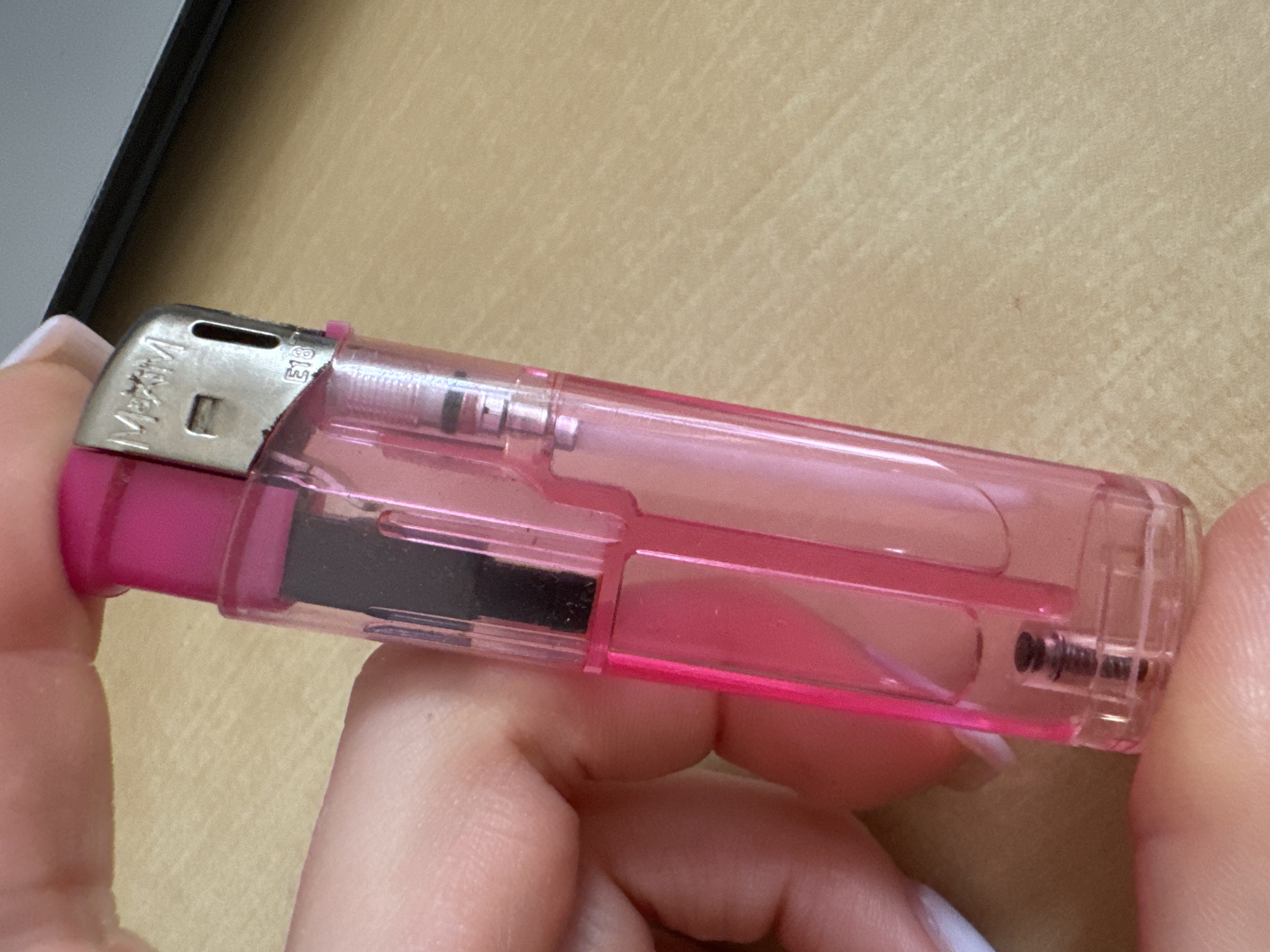 A close-up, eye-level shot focuses on a hand holding a pink, translucent plastic lighter. The lighter has a metallic top with the brand name "Maxim" visible. A small code "E13" is also present on the metallic part. The hand appears to be that of an adult with neatly manicured nails, possibly painted white. The lighter is positioned vertically, with the thumb and forefinger holding the body. The background is a light brown, textured surface, possibly a wooden desk or table. The lighting is neutral, casting soft shadows. The scene is devoid of any elements that would suggest a specific location within the city of Murcia, Spain, or any other urban environment.